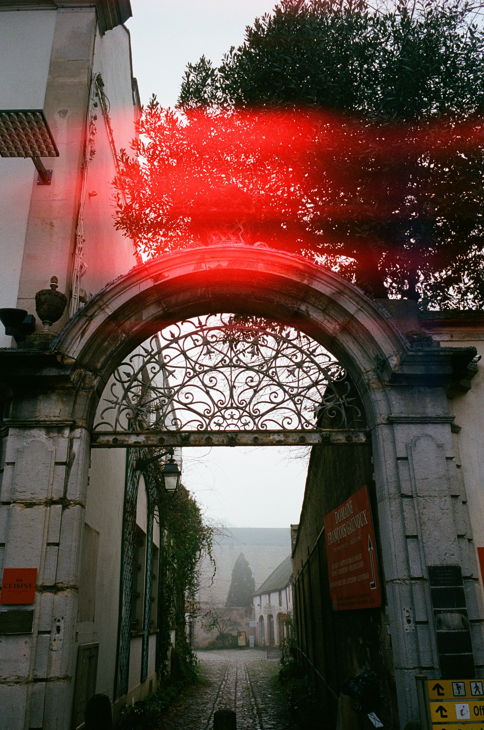 Une porte en pierre avec une grille en fer forgé, menant à une cour pavée avec des bâtiments blancs, un arbre et un ciel brumeux.