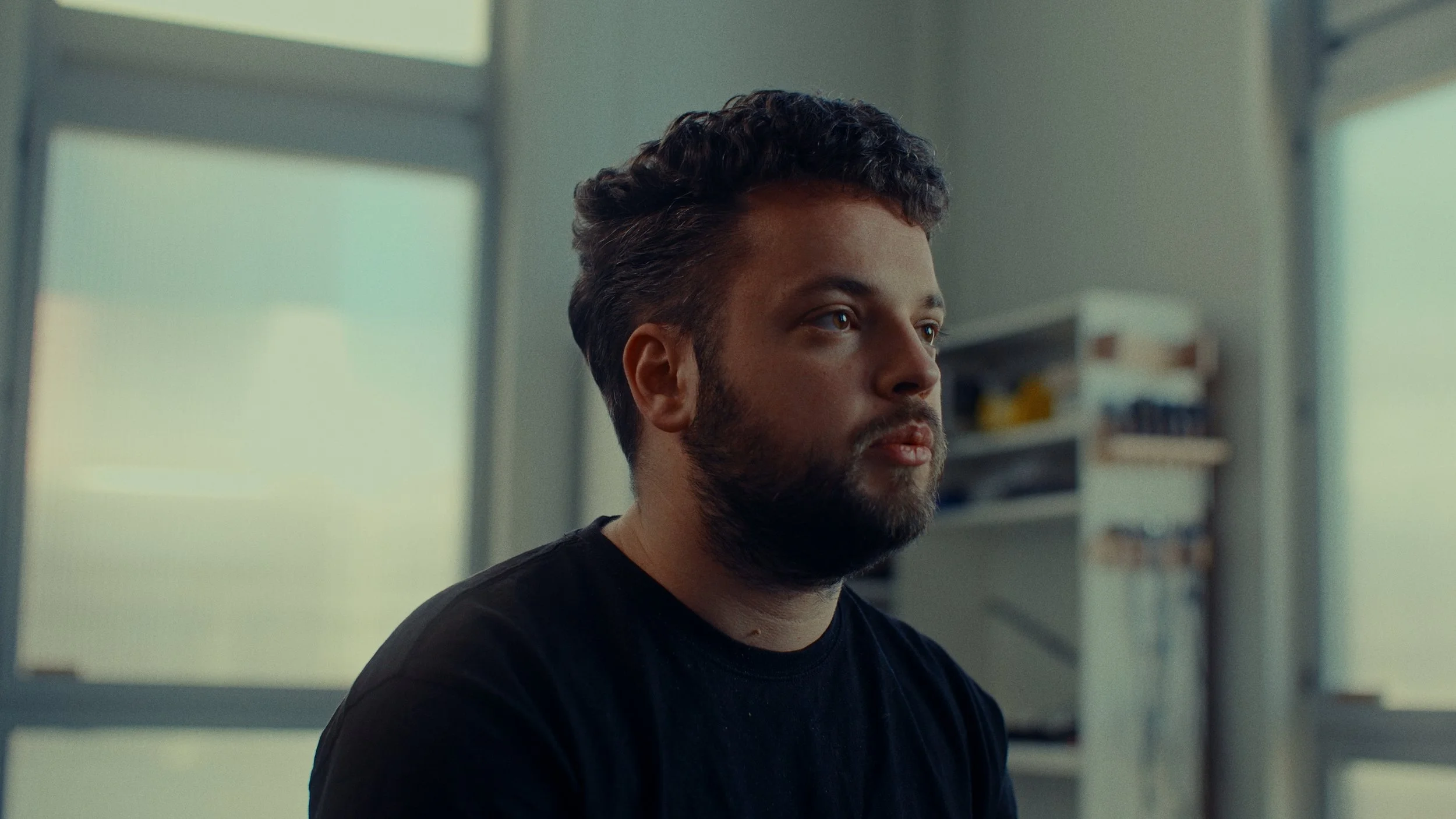 Homme avec barbe en intérieur, regardant à droite, près d'une fenêtre