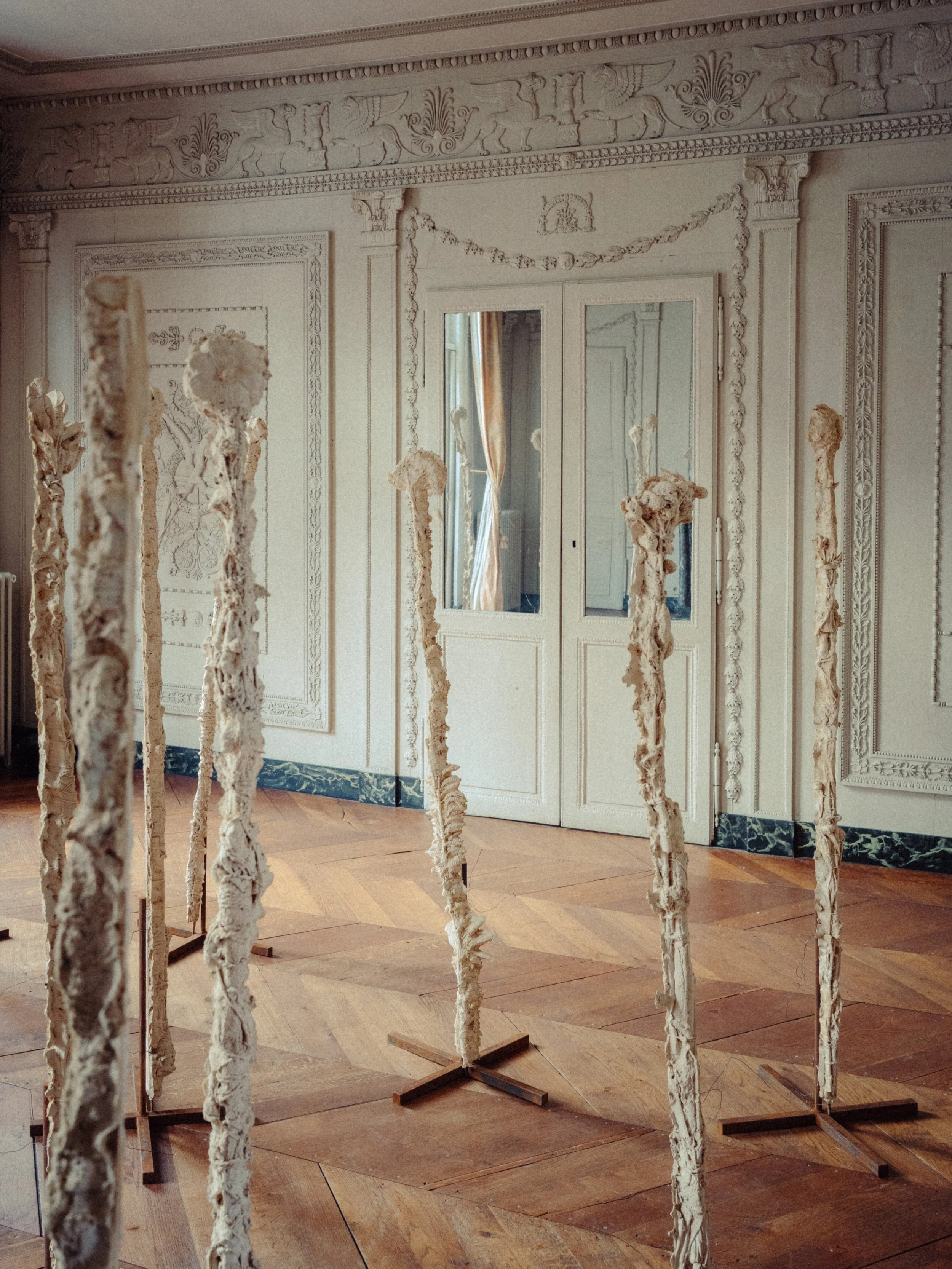 Salle ornée avec des moulures ornementales sur les murs et le plafond, présentant plusieurs sculptures en matériaux légers suspendues sur des supports, avec des fenêtres laissant entrer la lumière naturelle.
