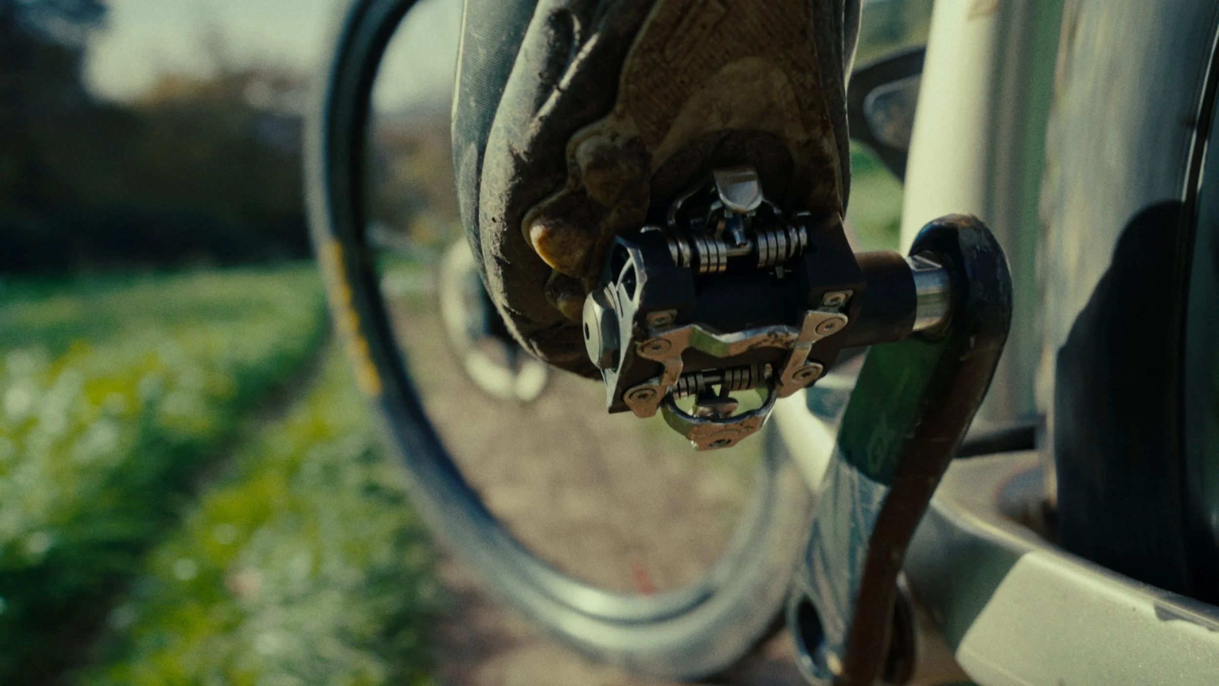 Détail d'une pédale de vélo attachée à la boîte de vitesses d'une bicyclette, avec un gant beige sale. Arrière-plan flou avec un chemin et de l'herbe verte.