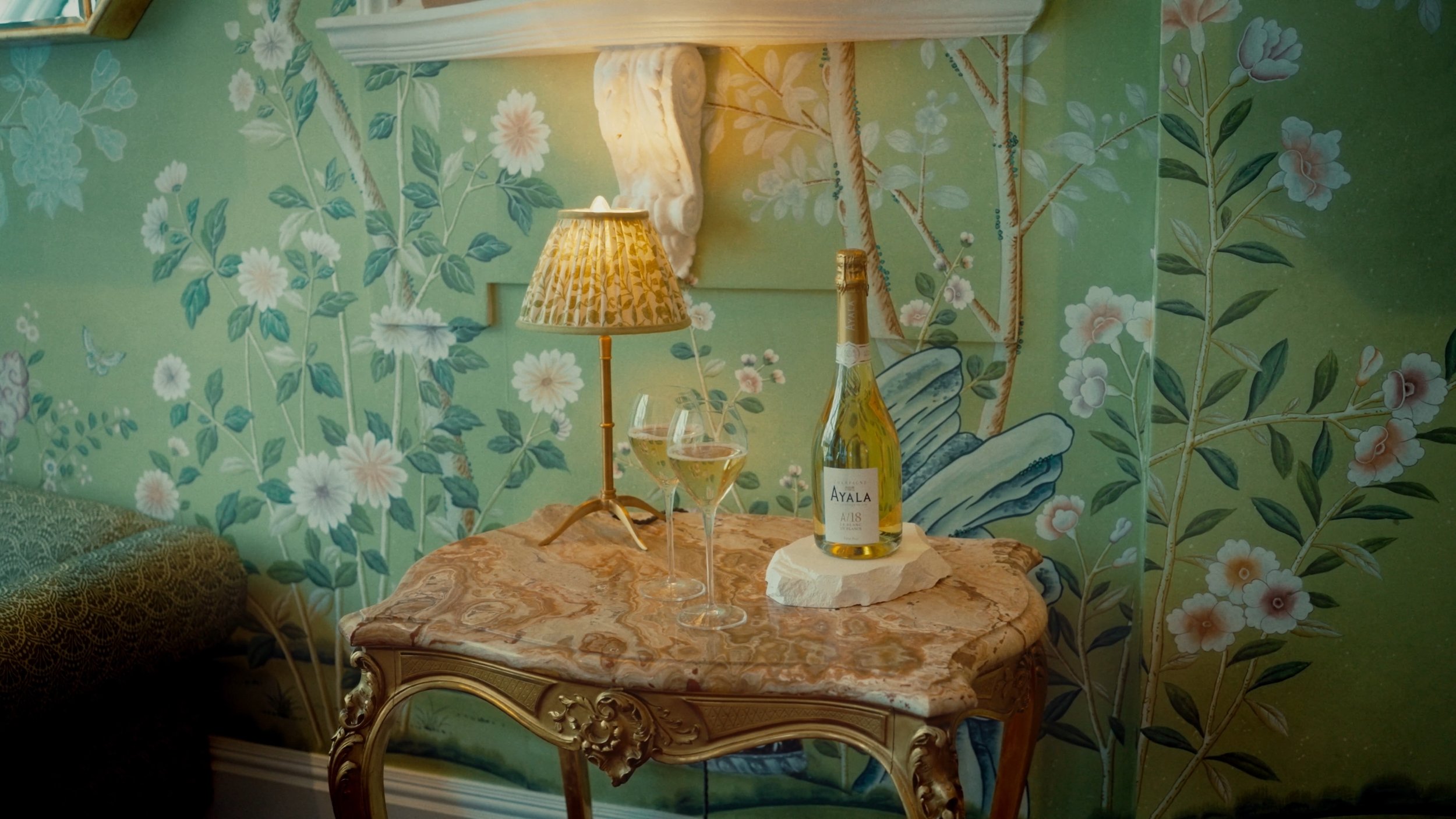 Table élégante en marbre avec une bouteille de vin, deux verres de vin blanc et une lampe sur un fond de mur peint avec des fleurs et des branches vertes.