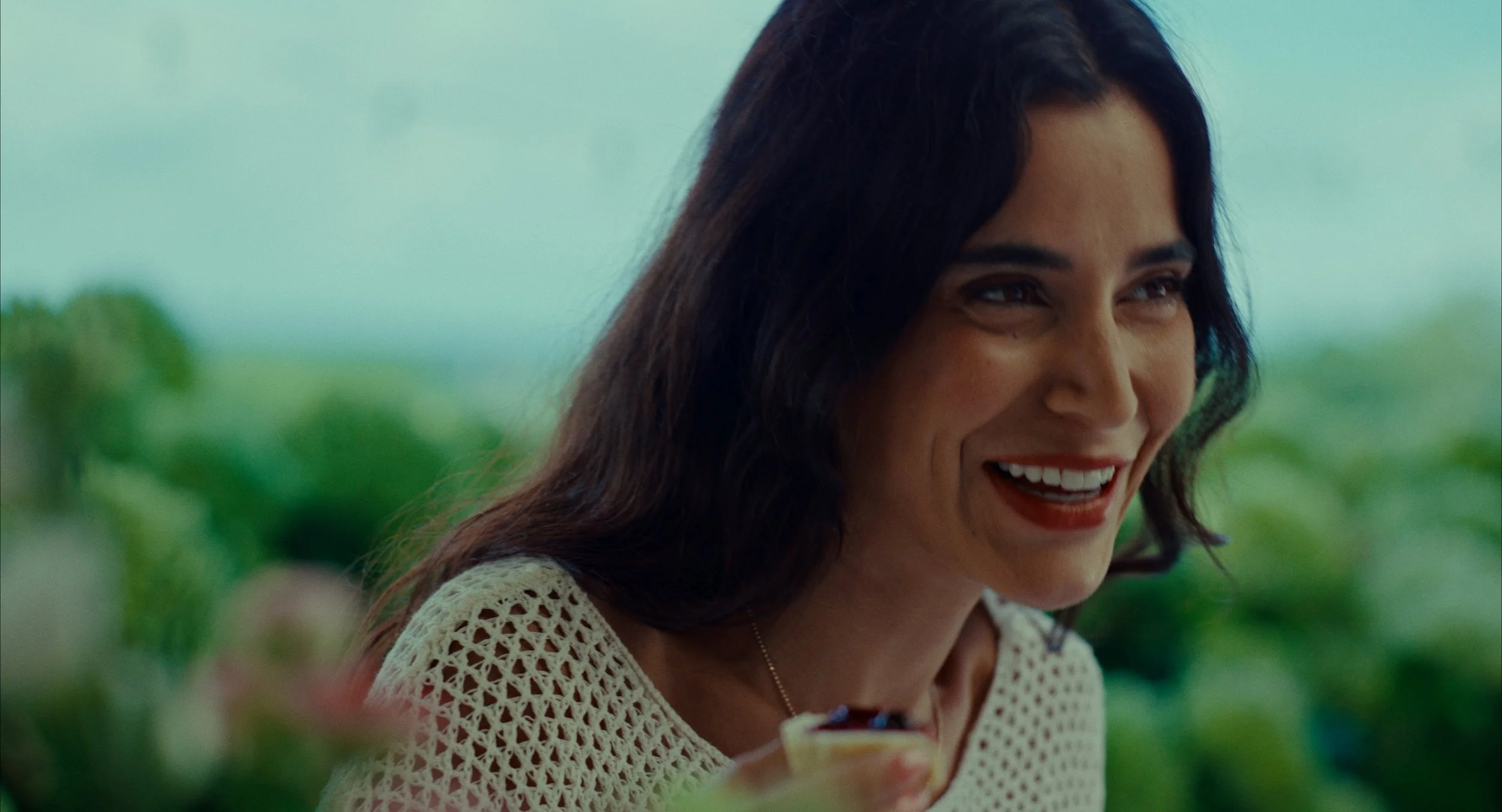 Une femme souriante avec des cheveux longs et noirs, portant un vêtement en crochet beige, tenant une tasse, devant un arrière-plan naturel flou.