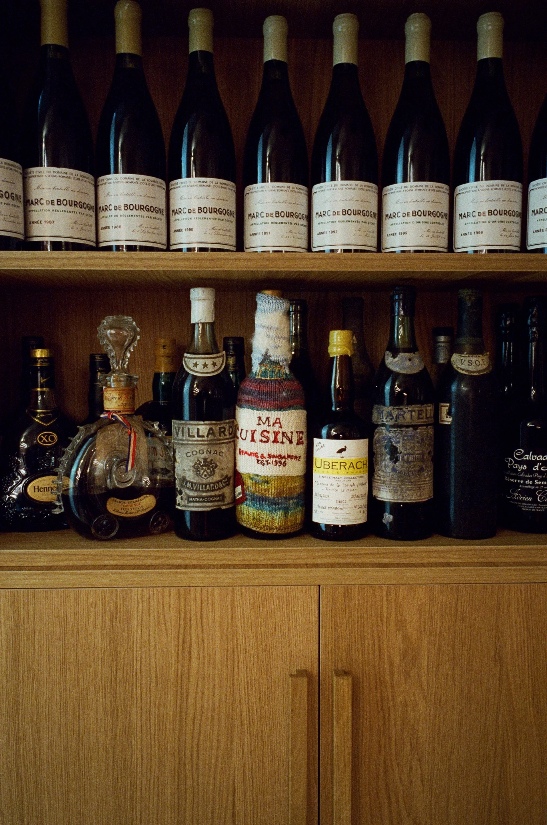 Étagère en bois avec des bouteilles de vin et de liqueur, dont plusieurs bouteilles de Marc de Bourgogne sur la partie supérieure, et diverses autres bouteilles d'alcool, dont un cognac et une bouteille portant un étui en laine, sur la partie inférie