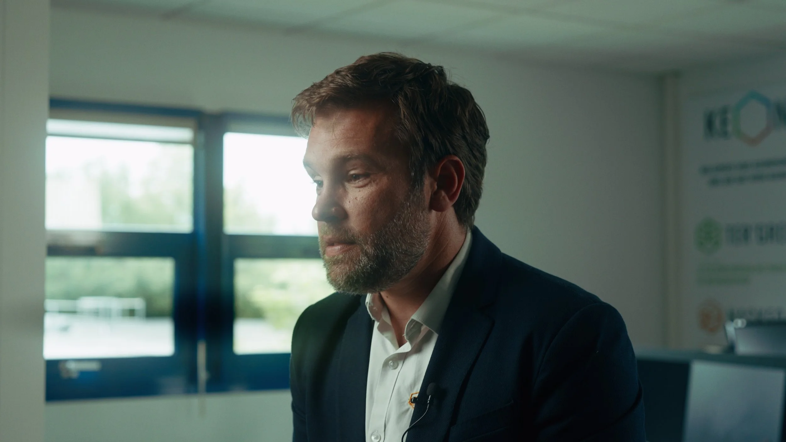 Un homme avec une barbe et des cheveux bruns, habillé en veste sombre et chemise blanche, semble pensif dans une pièce avec de grandes fenêtres en arrière-plan.