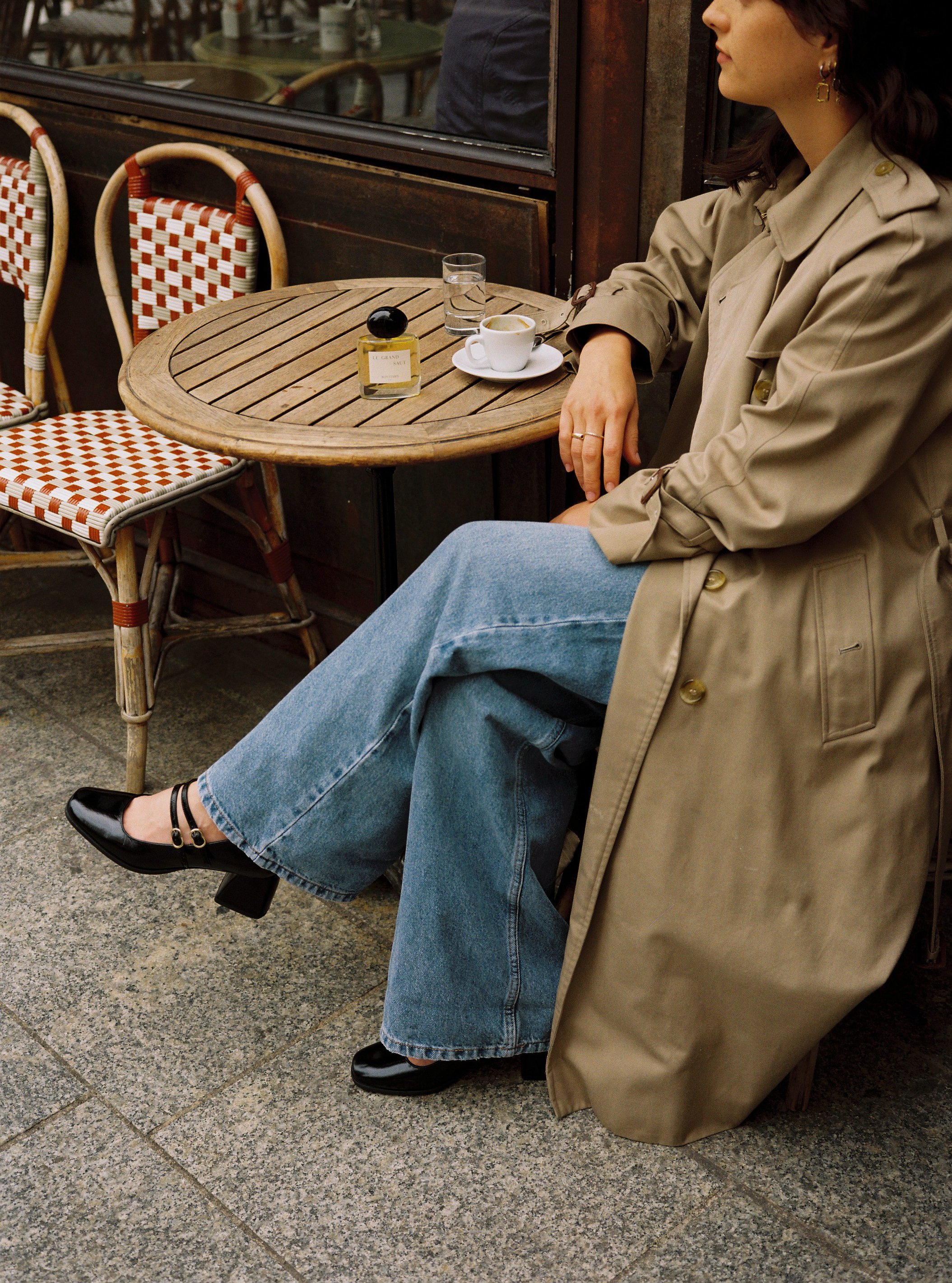 Une femme assise à une table en terrasse de café, portant un trench beige, un jean large, et des chaussures noires avec talon. Sur la table, il y a un verre d'eau, une tasse à café, et un flacon de parfum.