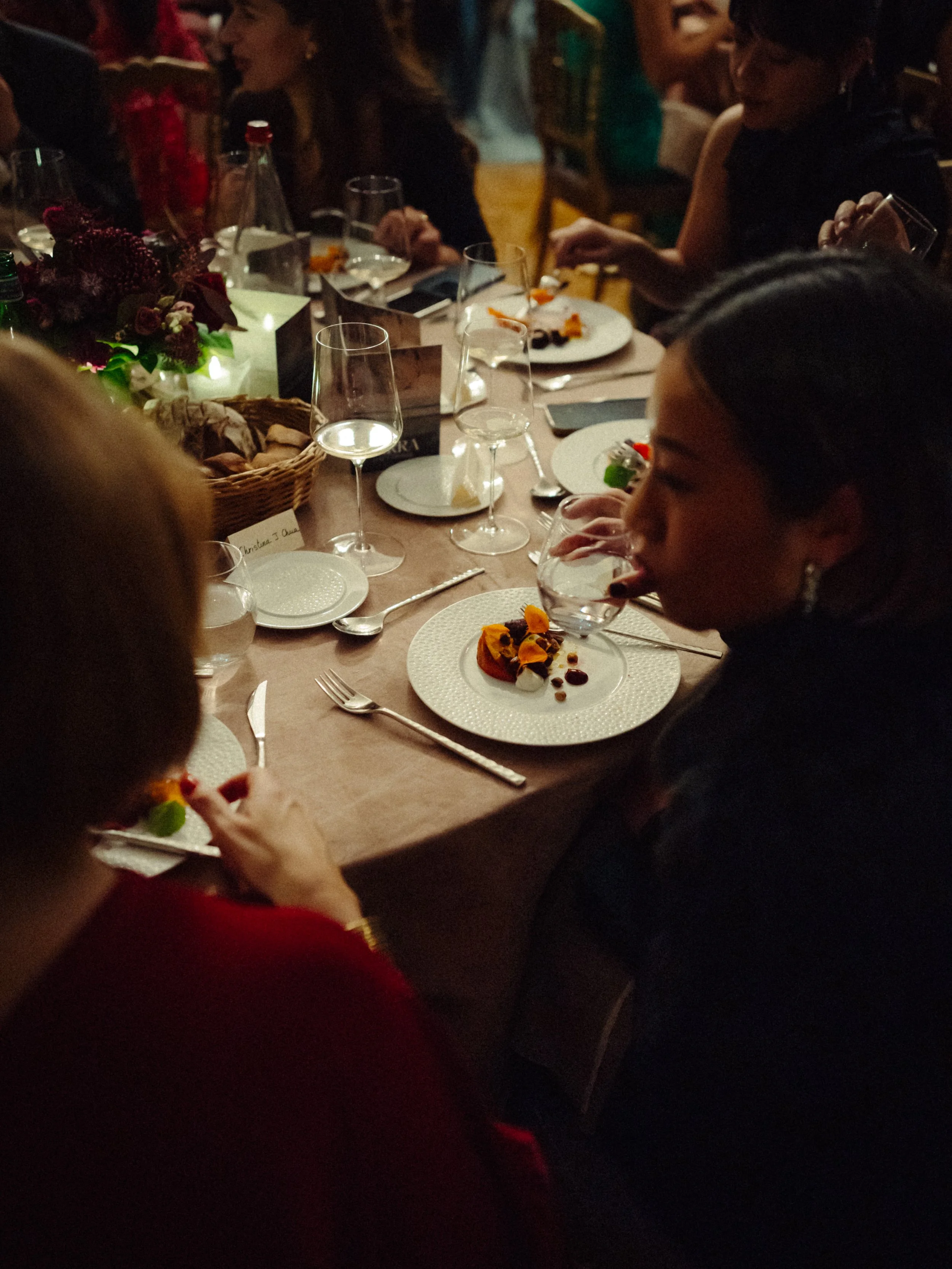 Groupe de personnes dégustant un repas en soirée dans une ambiance chaleureuse, assises autour d'une table décorée avec des plats, des verres et des fleurs.