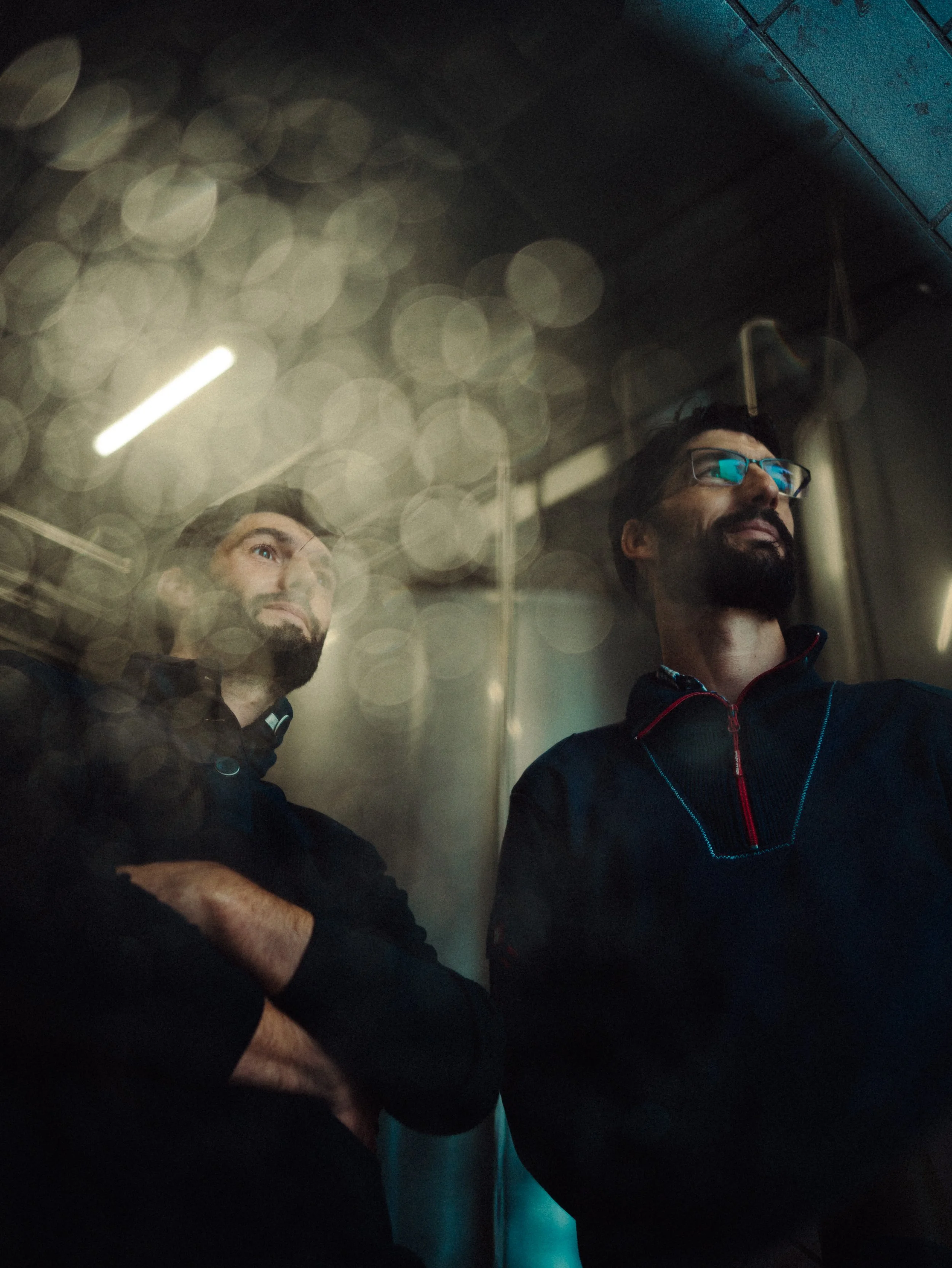 Deux hommes regardant à travers une vitre, avec des reflets flous et une lumière au plafond dans un environnement intérieur.