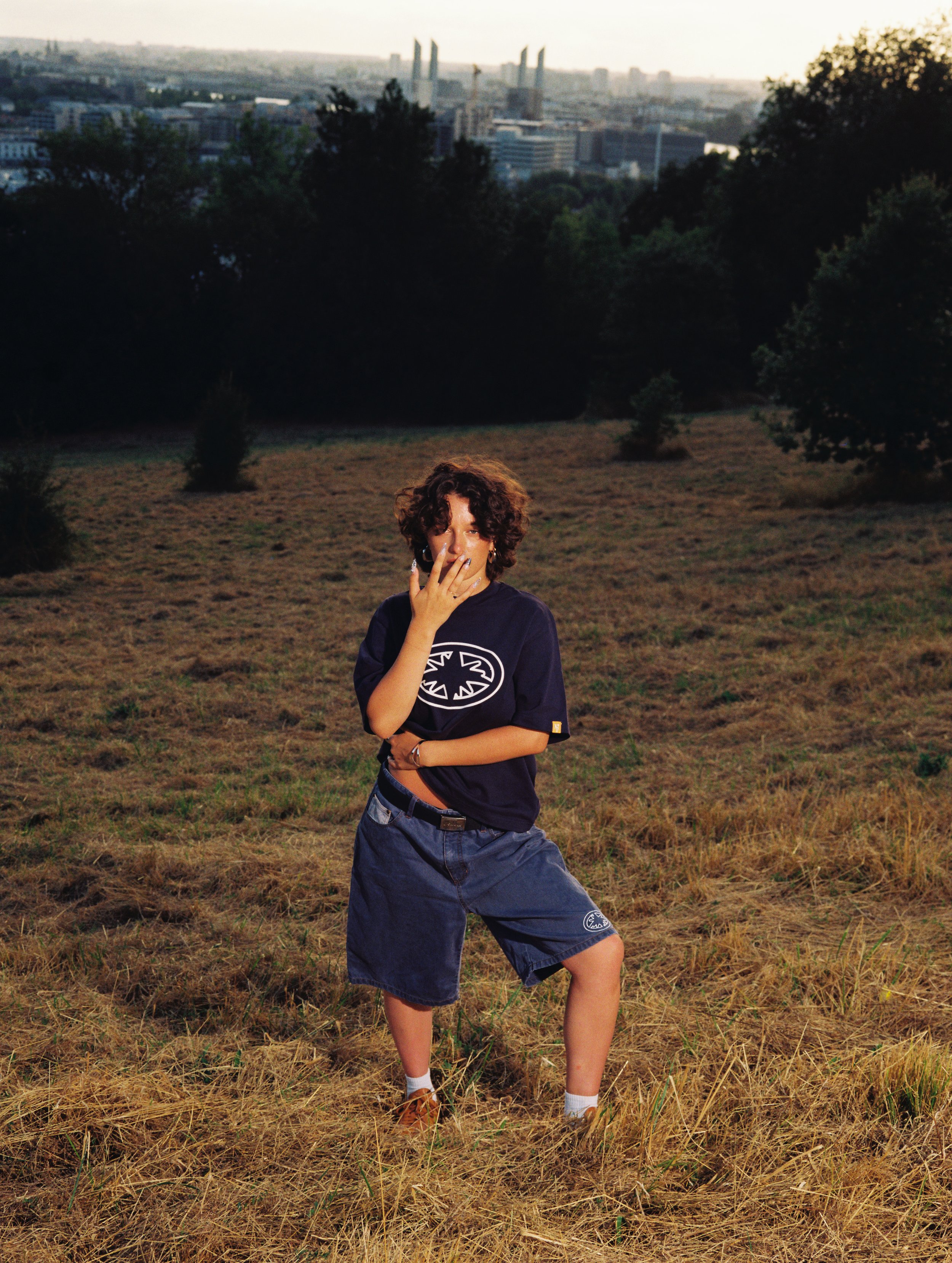 Une personne debout dans un champ, portant un t-shirt noir, un short bleu, et des chaussures marron, avec une ville en arrière-plan.