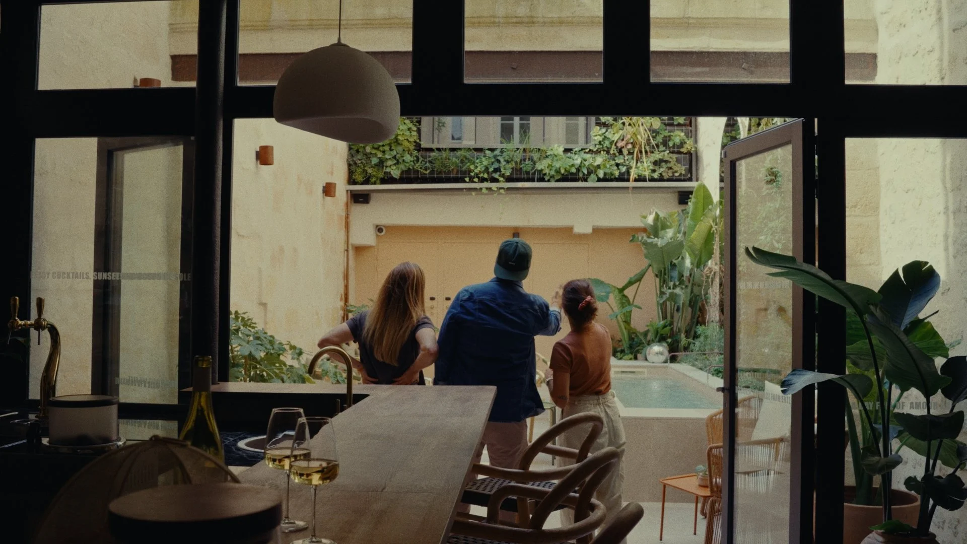 Vue d'une cuisine ouverte donnant sur une cour avec trois personnes debout près d'une piscine, vues depuis l'intérieur de la maison.