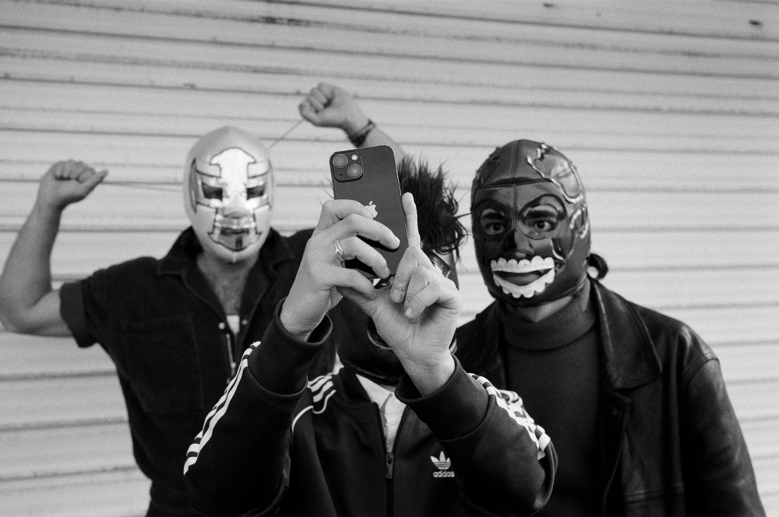 Trois personnes portant des masques de carnaval, une personne au centre prend une photo avec son téléphone, fond avec des volets en bois.