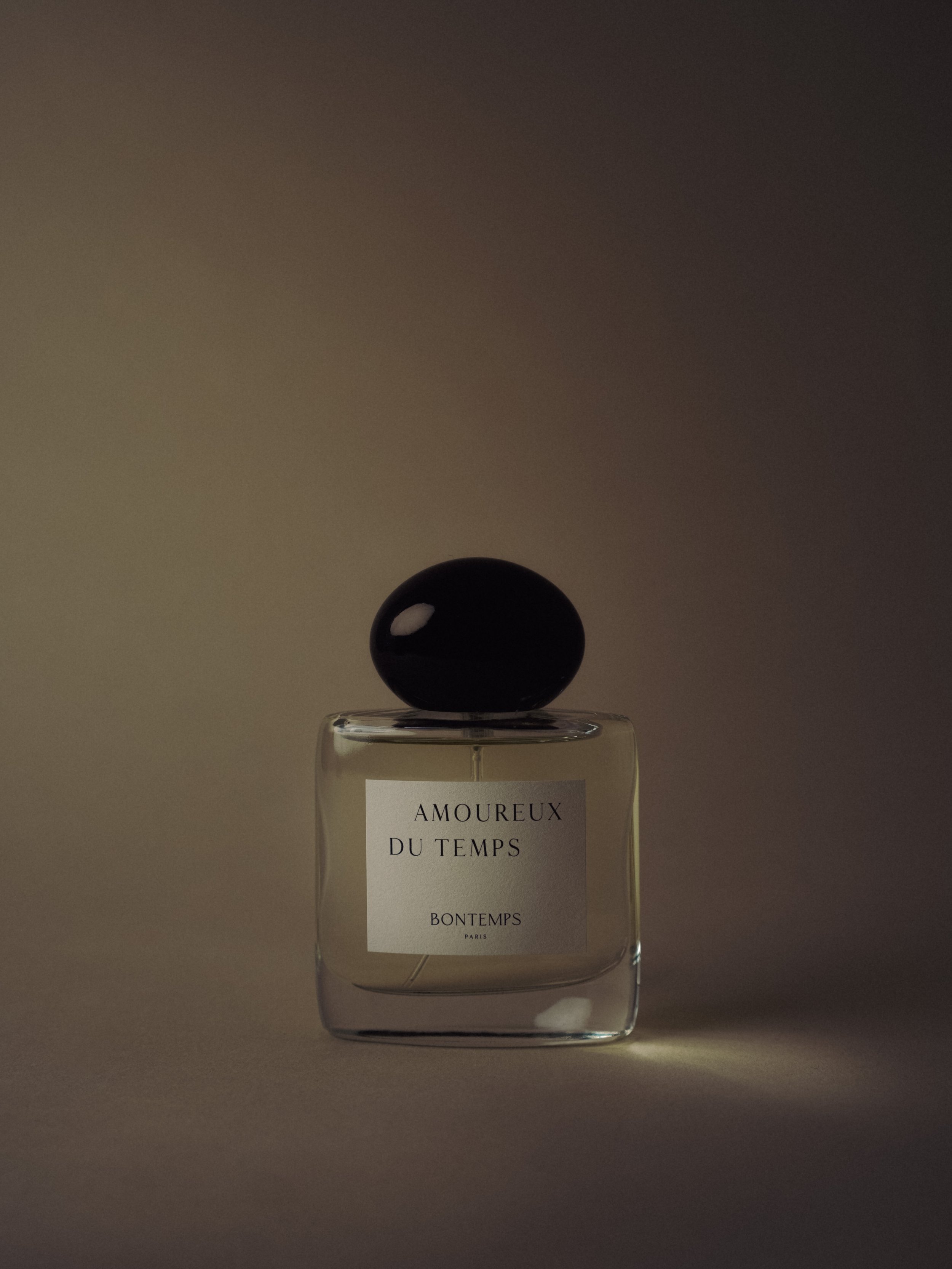 Flacon de parfum transparent avec un bouchon noir, étiquette blanche avec texte noir, placé sur une surface neutre avec un fond sombre et un éclairage doux.