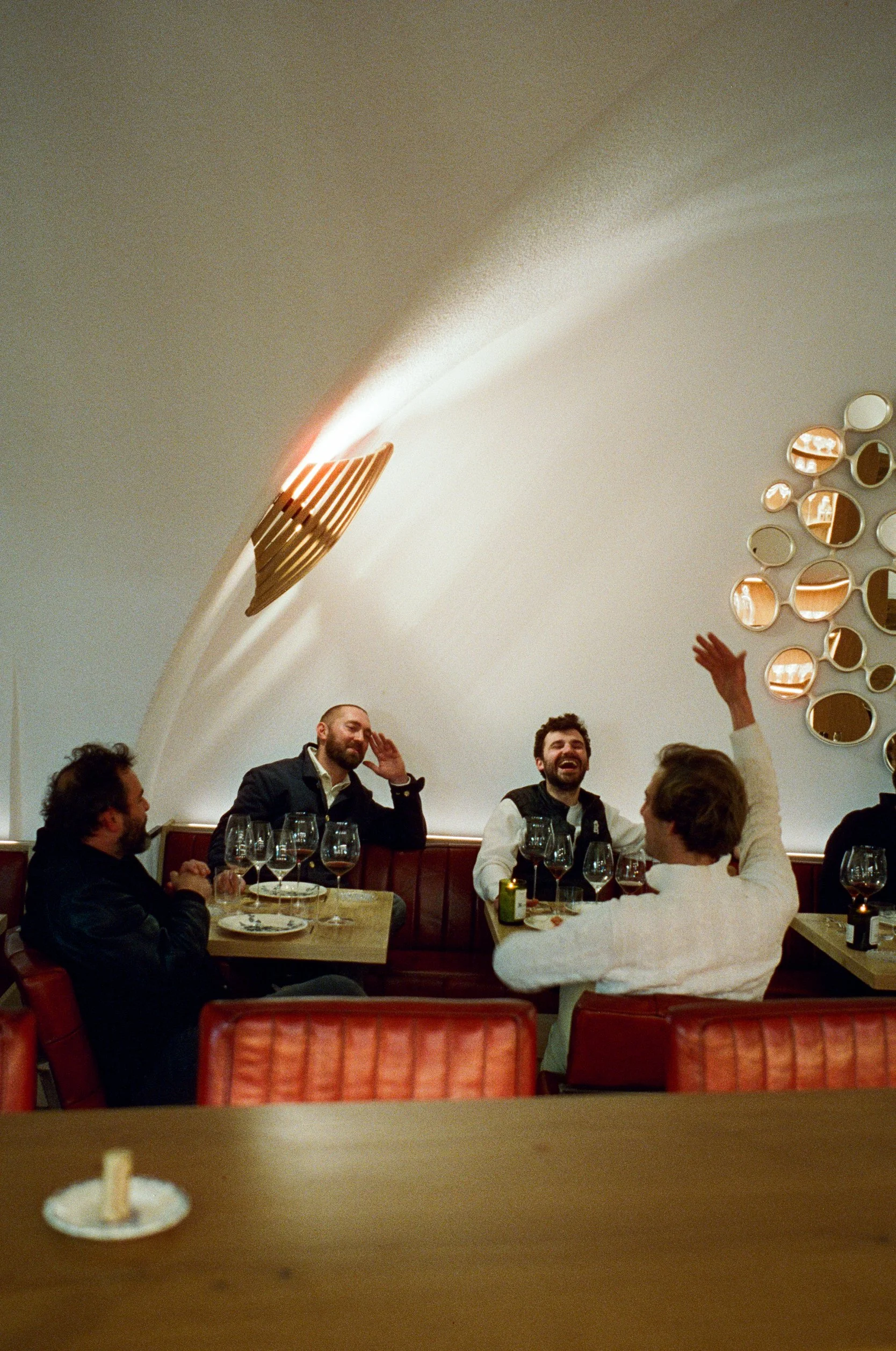 Quatre personnes rient et discutent dans un restaurant, l'un d'eux lève la main, avec des verres de vin sur la table et un mur décoratif avec des miroirs en boule en fond.