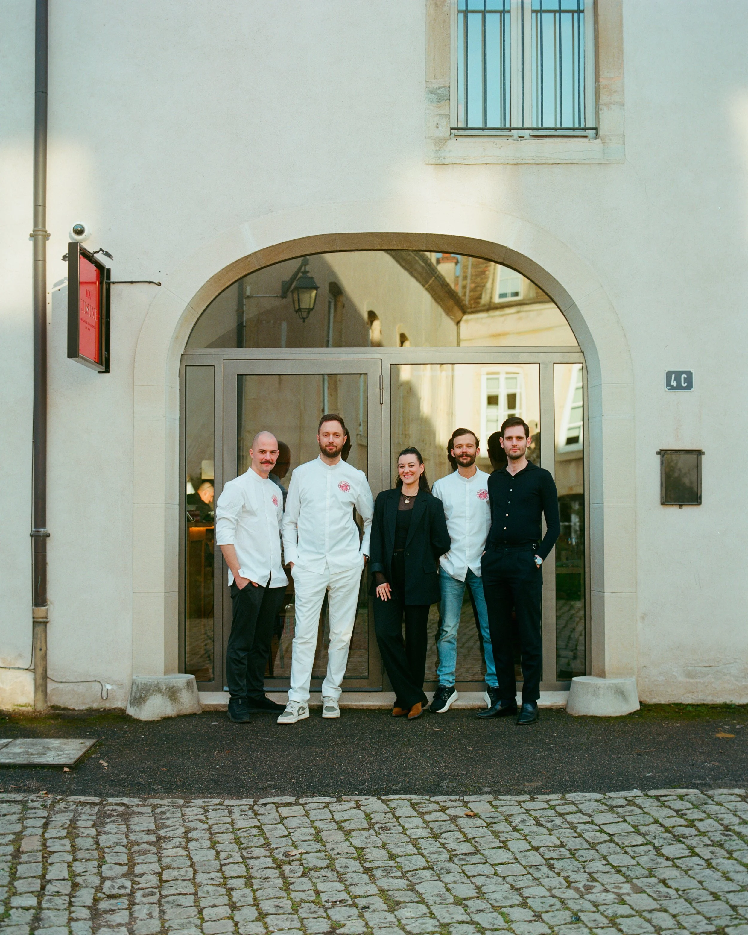 groupe de cinq personnes devant un restaurant avec une porte en verre, dont deux chefs en uniforme blanc et une femme avec un costume noir.