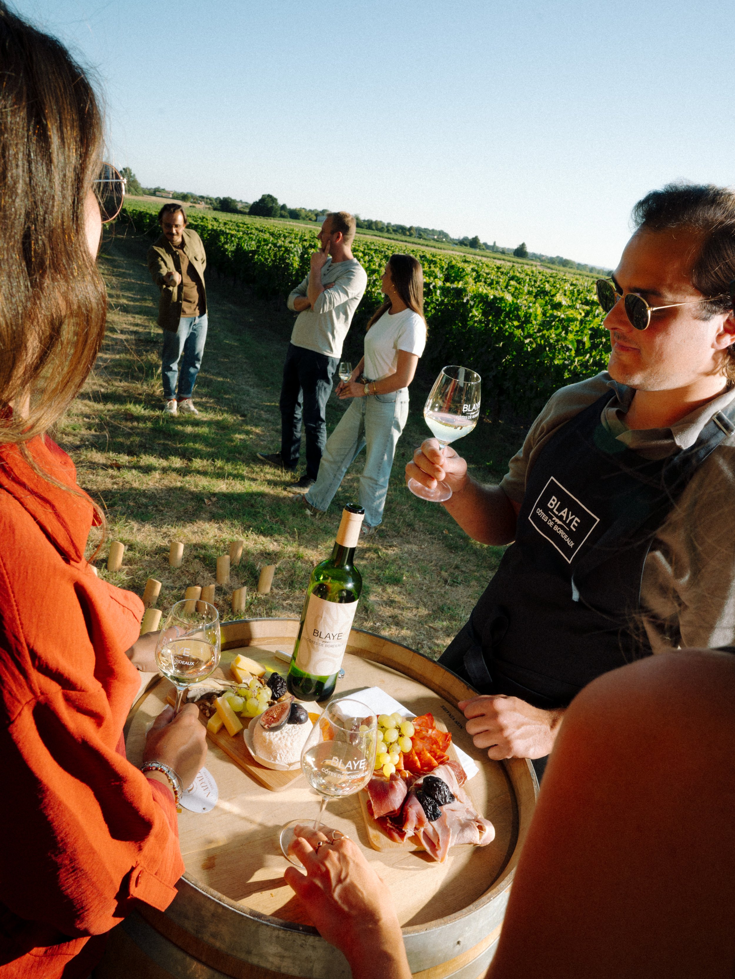 Un groupe de personnes dégustant du vin lors d'une dégustation en plein air dans une vigne avec un étalage de nourriture et de vin au centre.