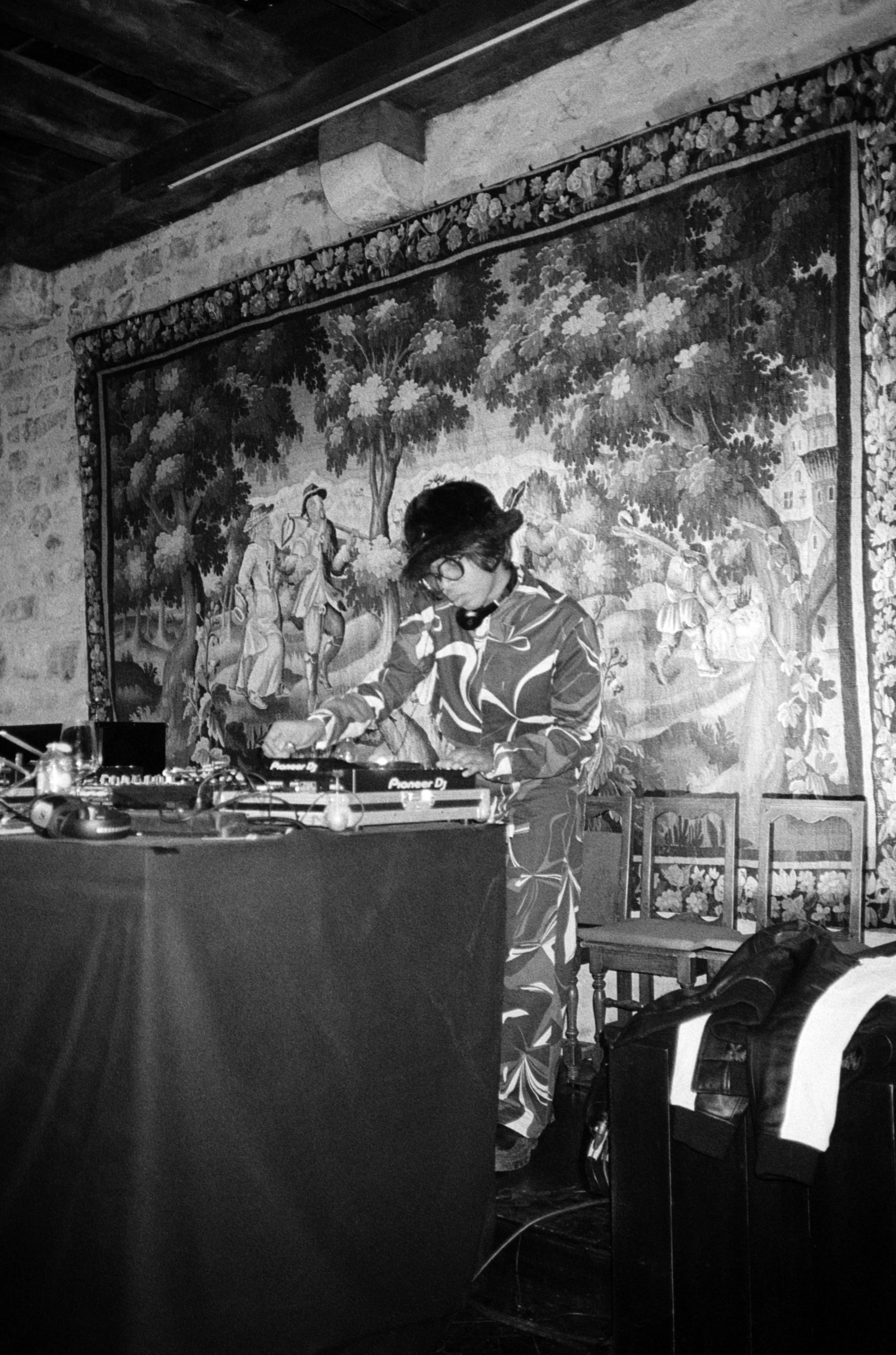 Une personne en tenue de DJ mixant avec un casque, devant un tableau ou une tapisserie illustrée d'un paysage avec des personnes dans la nature, dans une pièce aux murs en pierre.