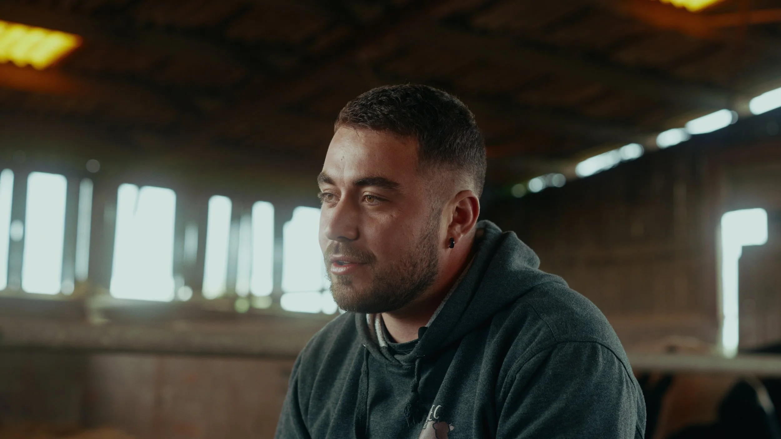 Jeune homme avec barbe, maquillée, portant un sweat gris, assis à l'intérieur d'un bâtiment en bois, avec des fenêtres lumineuses derrière lui.