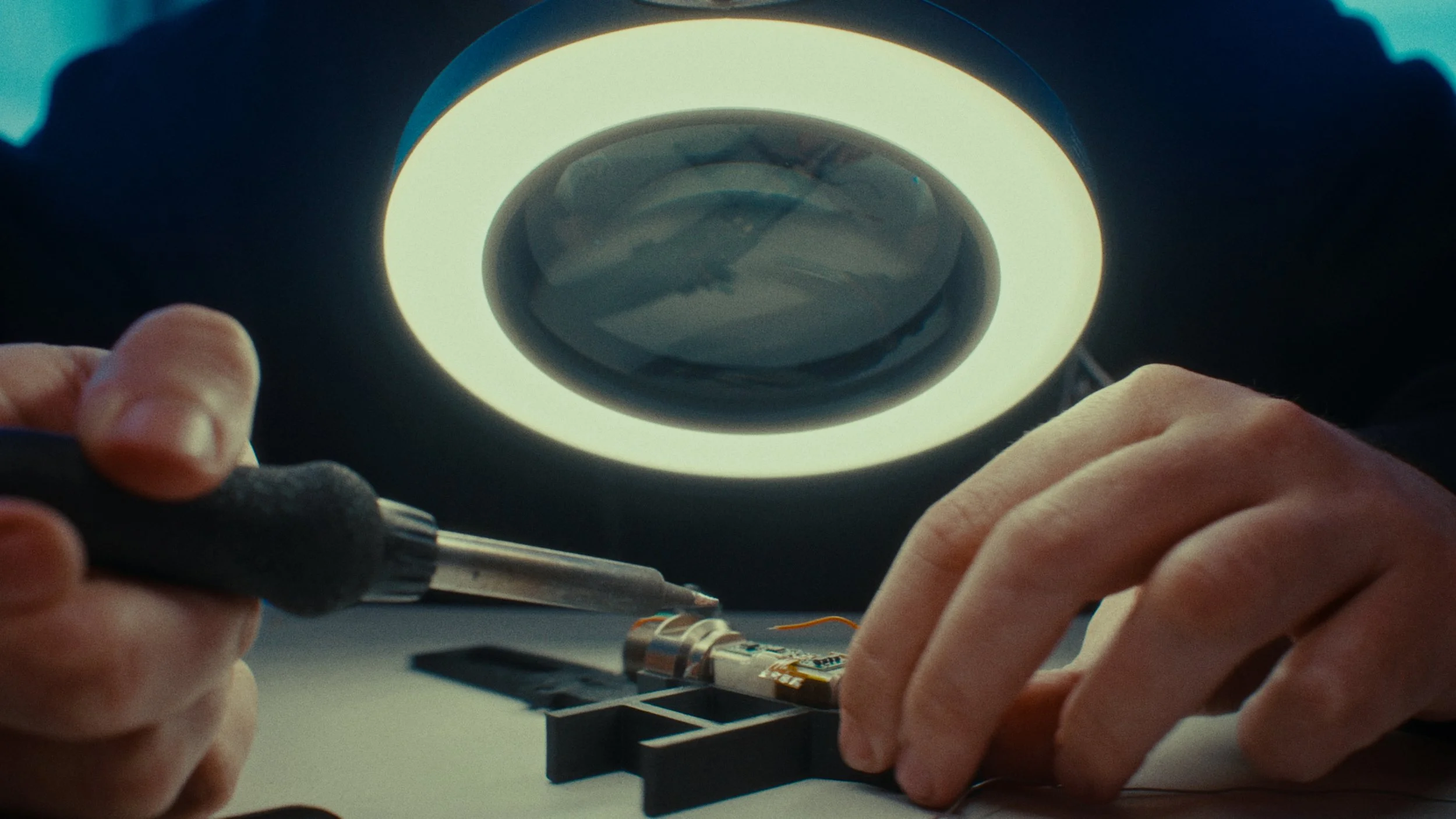 Un technicien effectue une réparation ou une inspection à l'aide d'un fer à souder sur un circuit imprimé sous une lumière de travail.