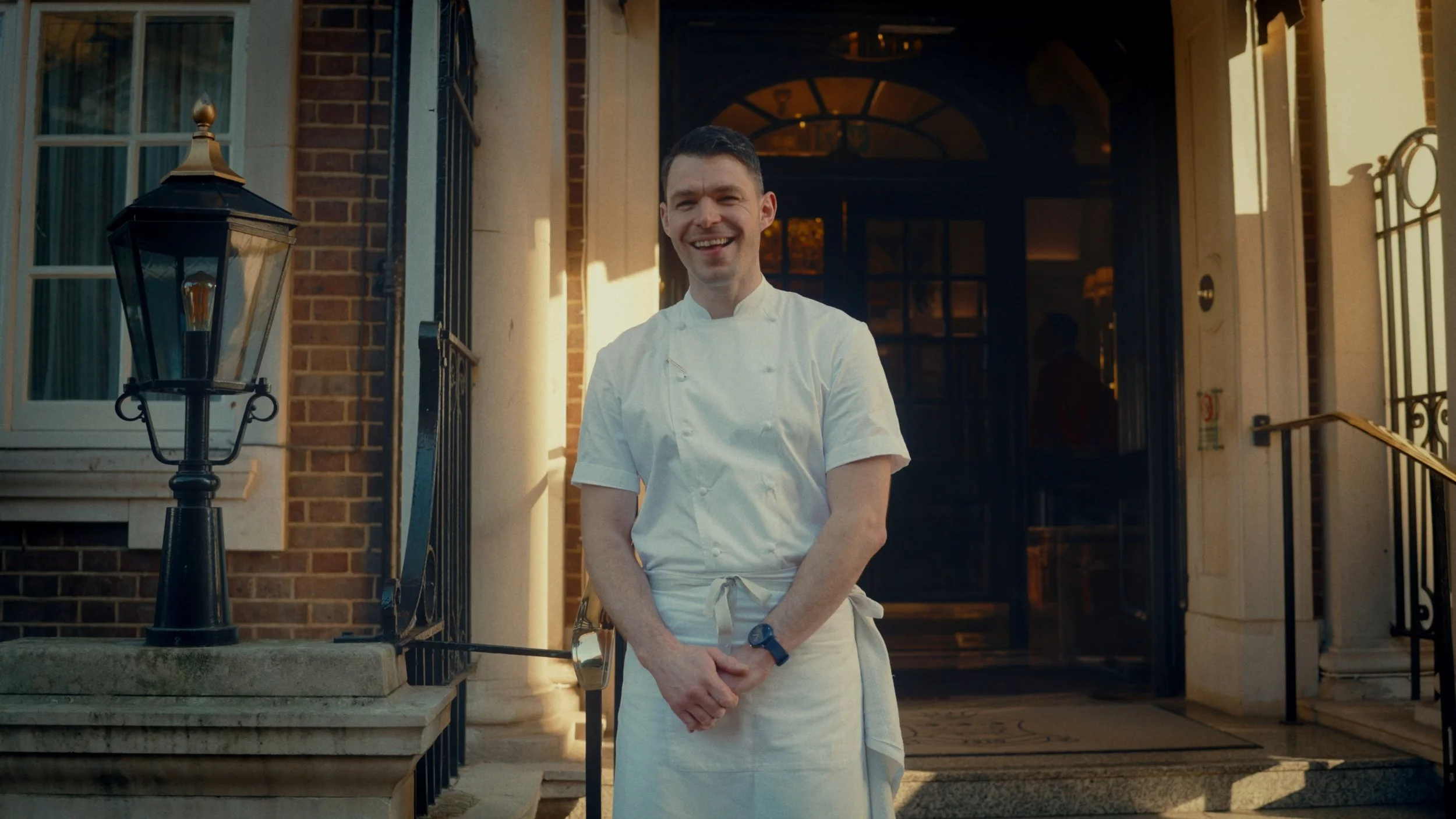 Un chef souriant en uniforme blanc, debout à l'entrée d'une maison ou d'un restaurant, avec un bâtiment en brique et un lampadaire à sa gauche.
