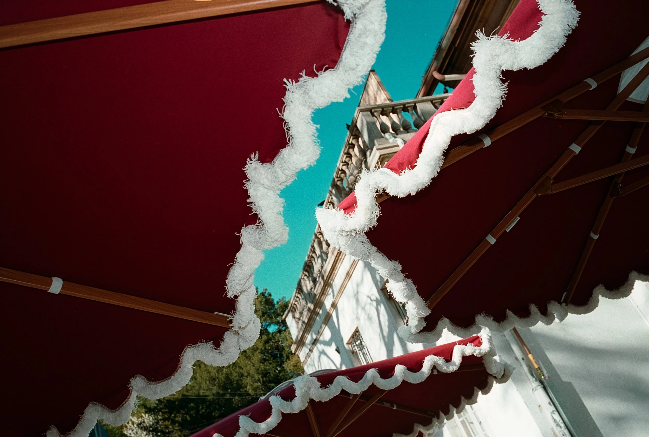 Tentes rouges avec bordures blanches en bord de mer ou de lieu public, avec un bâtiment blanc et un arbre vert en arrière-plan, sous un ciel bleu.