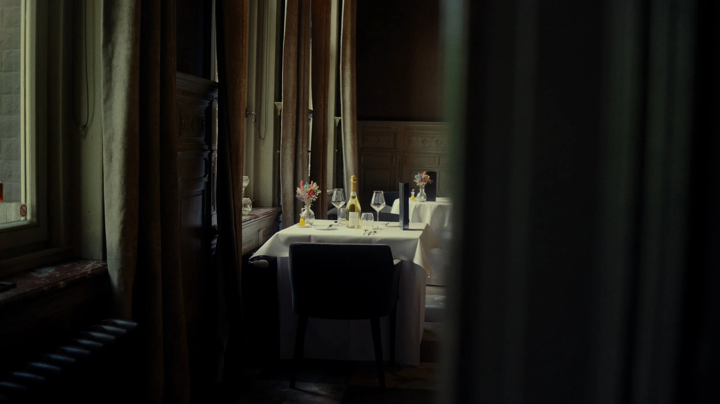 Une salle à manger élégante avec une table dressée de nappes blanches, des verres à vin, une bouteille de champagne, des bouquets de fleurs et des décorations, vue partiellement à travers des rideaux.
