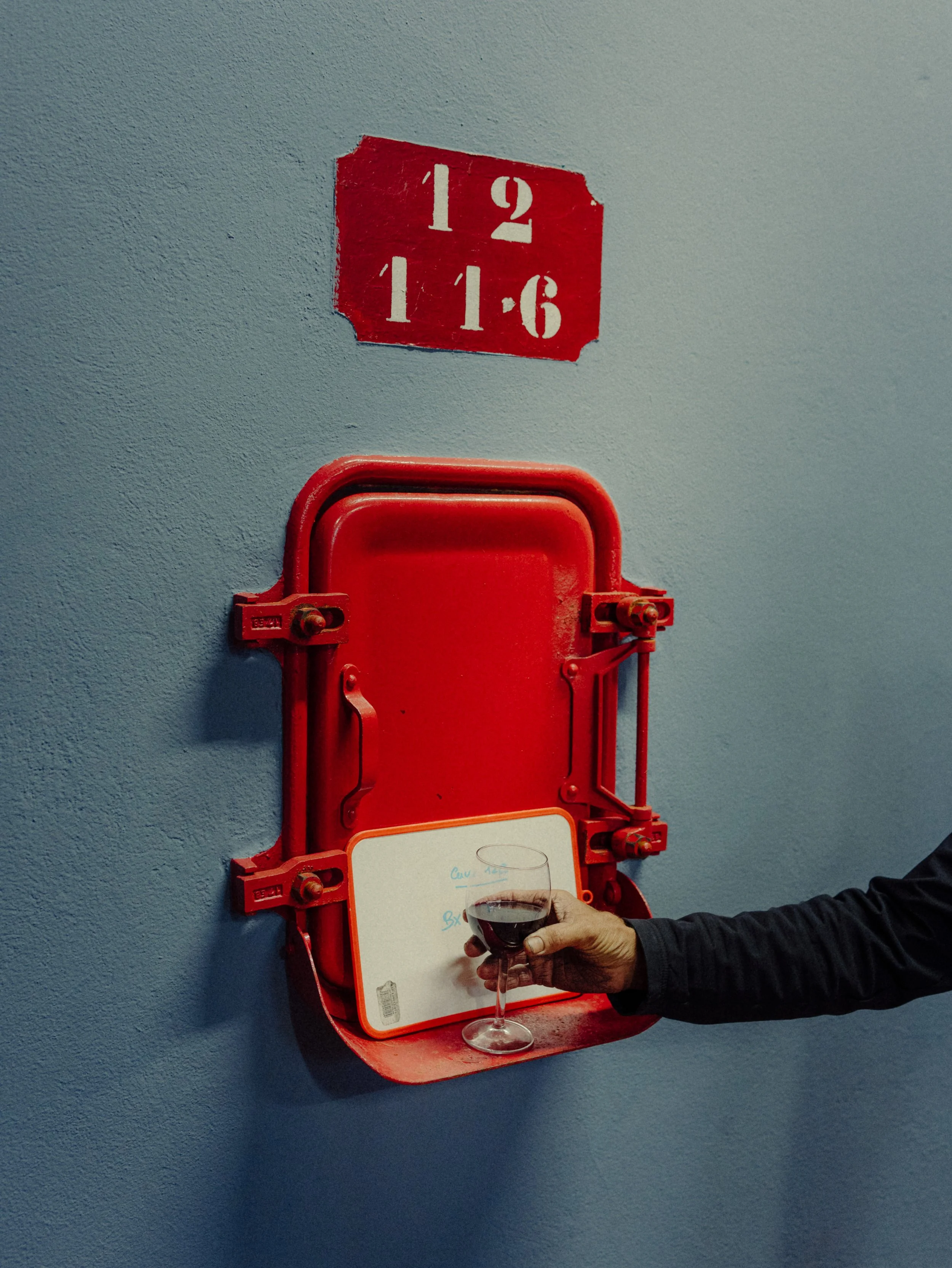 Une main tenant un verre de vin rouge devant une boîte à vin rouge fixée au mur, avec un tableau blanc dessous. Le mur a une inscription en rouge avec le texte '12 11-6'.