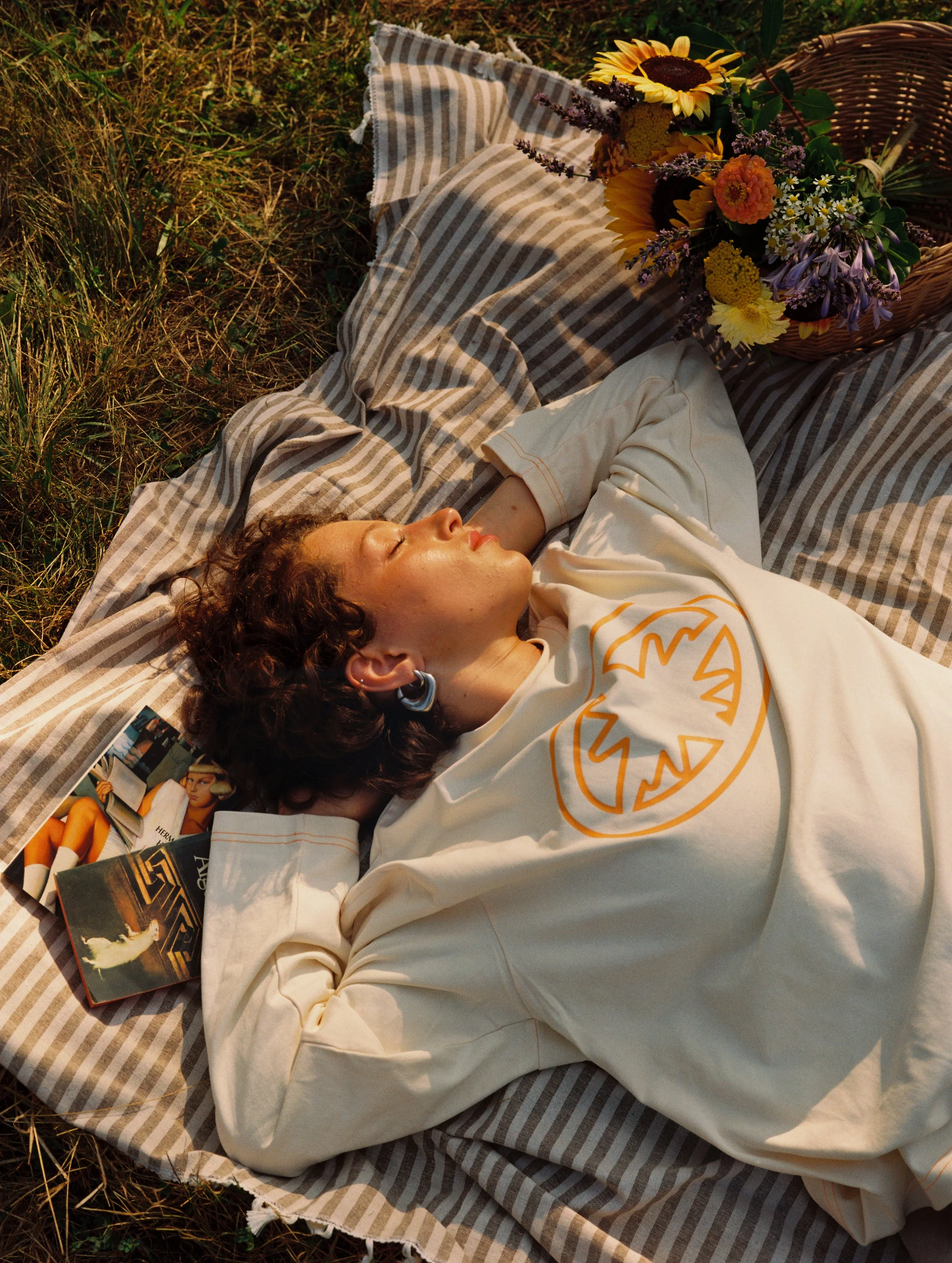 Une femme allongée sur une couverture rayée, avec des fleurs dans un panier à proximité, semble se détendre à l'extérieur à la lumière du soleil.