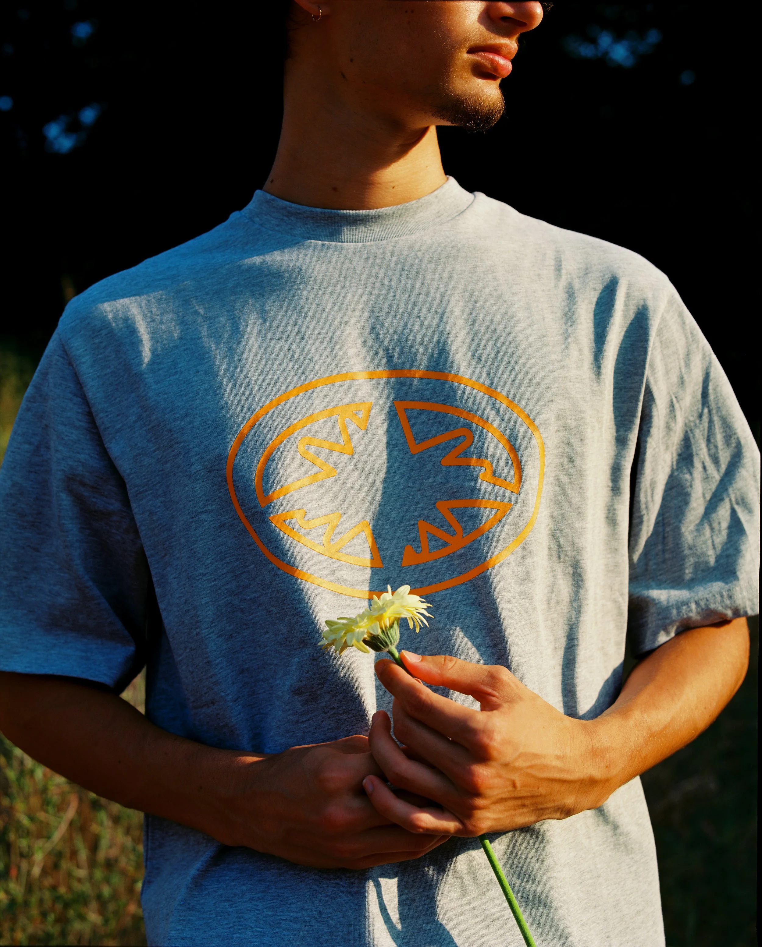 Une personne portant un t-shirt gris avec un logo orange et tenant une fleur blanche dans ses mains.