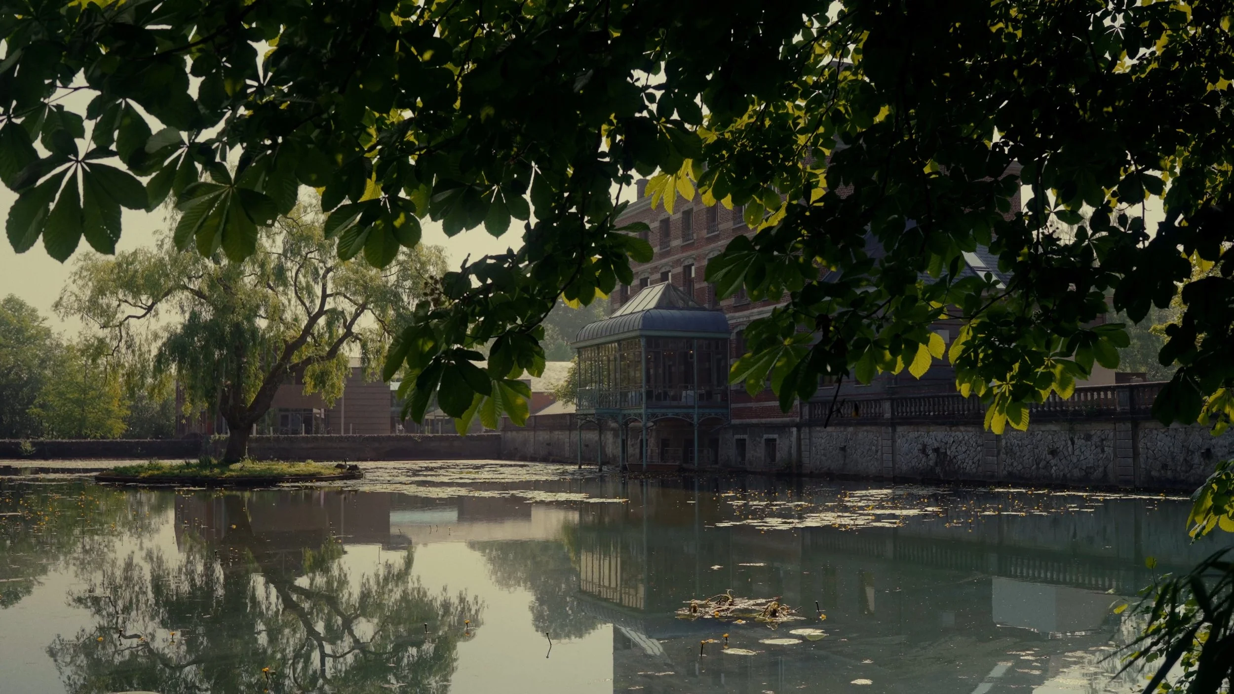 Une scène paisible avec un étang entouré d'arbres verdoyants, un bâtiment ancien en pierre avec une verrière en métal et une terrasse, le tout reflété dans l'eau calme de l'étang.