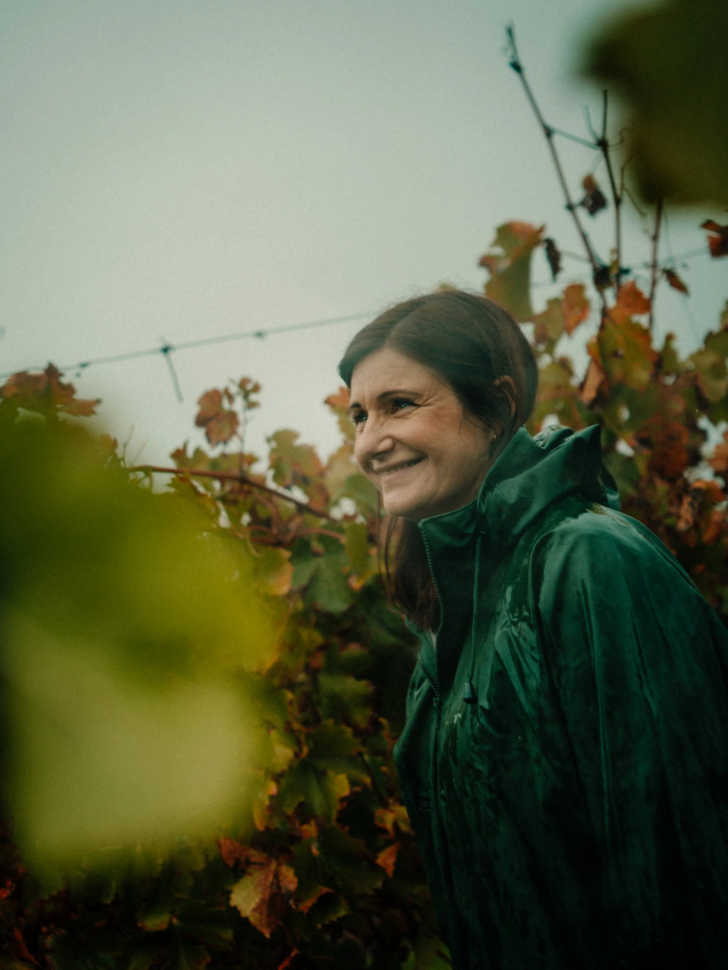 Une femme souriante portant un manteau vert foncé, dans un vignoble au moment de l'automne.