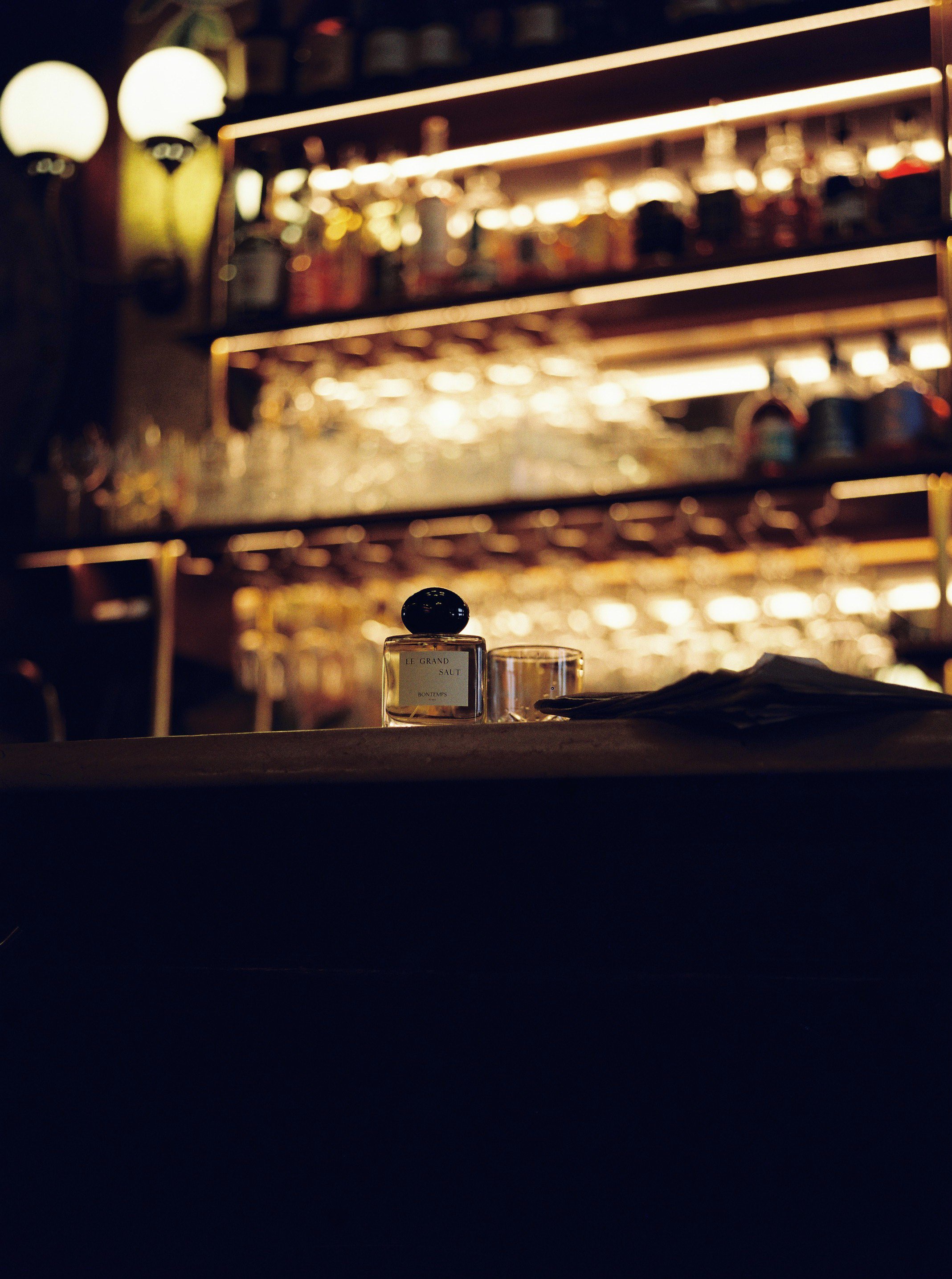 Une bouteille de parfum, un verre vide et un objet en bois avec un tissu noir sur un comptoir dans un bar ou un restaurant, avec des étagères éclairées en arrière-plan contenant divers bouteillons