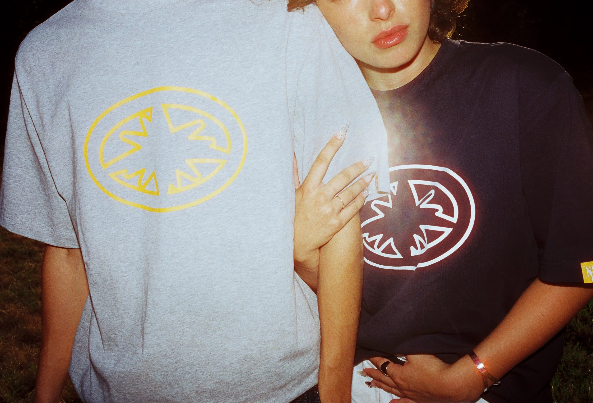 Deux personnes portant des t-shirts noirs et gris avec un logo circulaire jaune et blanc, côte à côte, dans un cadre extérieur la nuit.