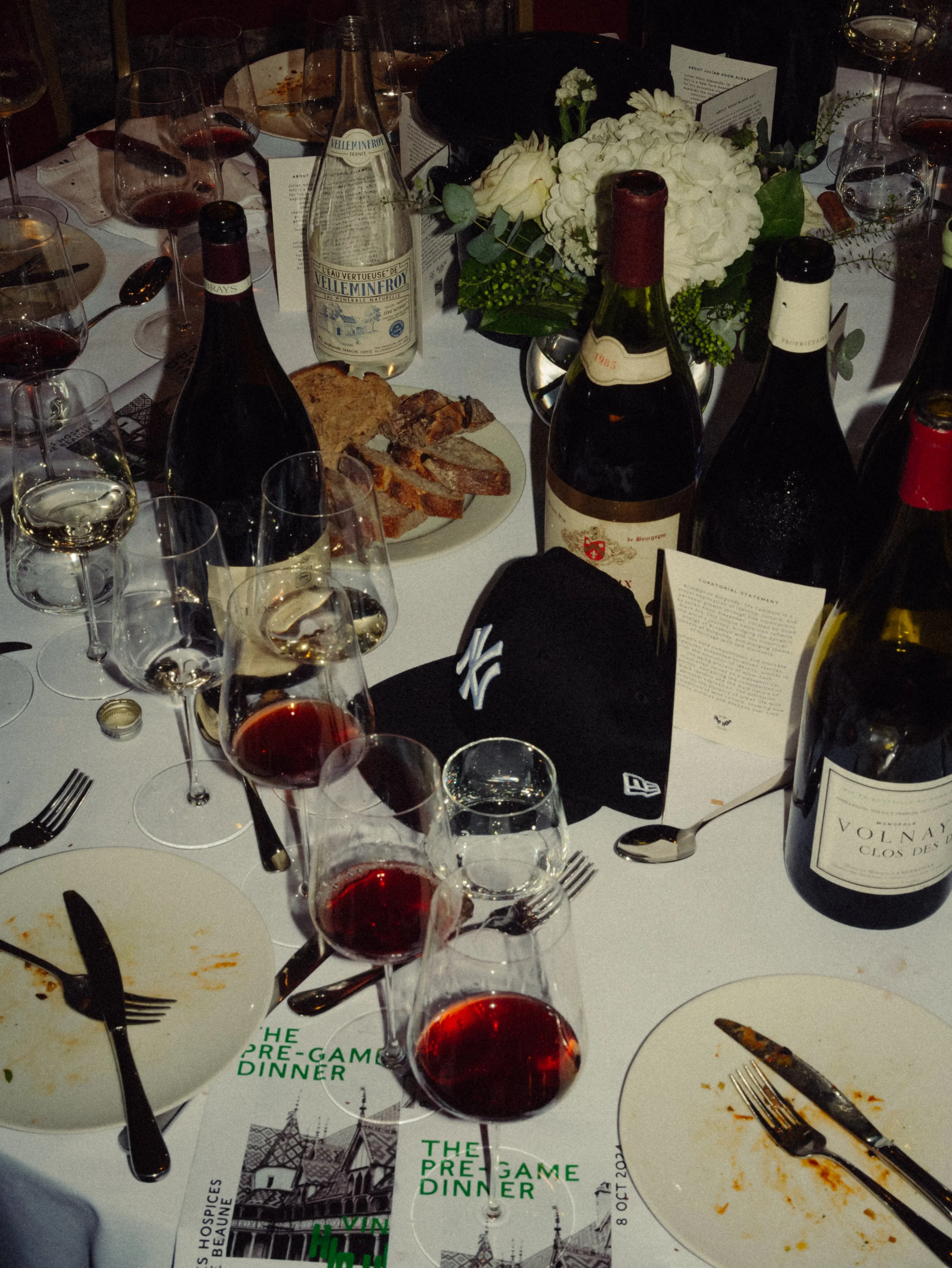 Table dressée pour un dîner avec plusieurs verres de vin rouge, une bouteille d'eau minérale, et des bouteilles de vin rouge, accompagnée d'un bouquet de fleurs blanches en centre de table. Il y a aussi des assiettes avec de la nourriture partielleme