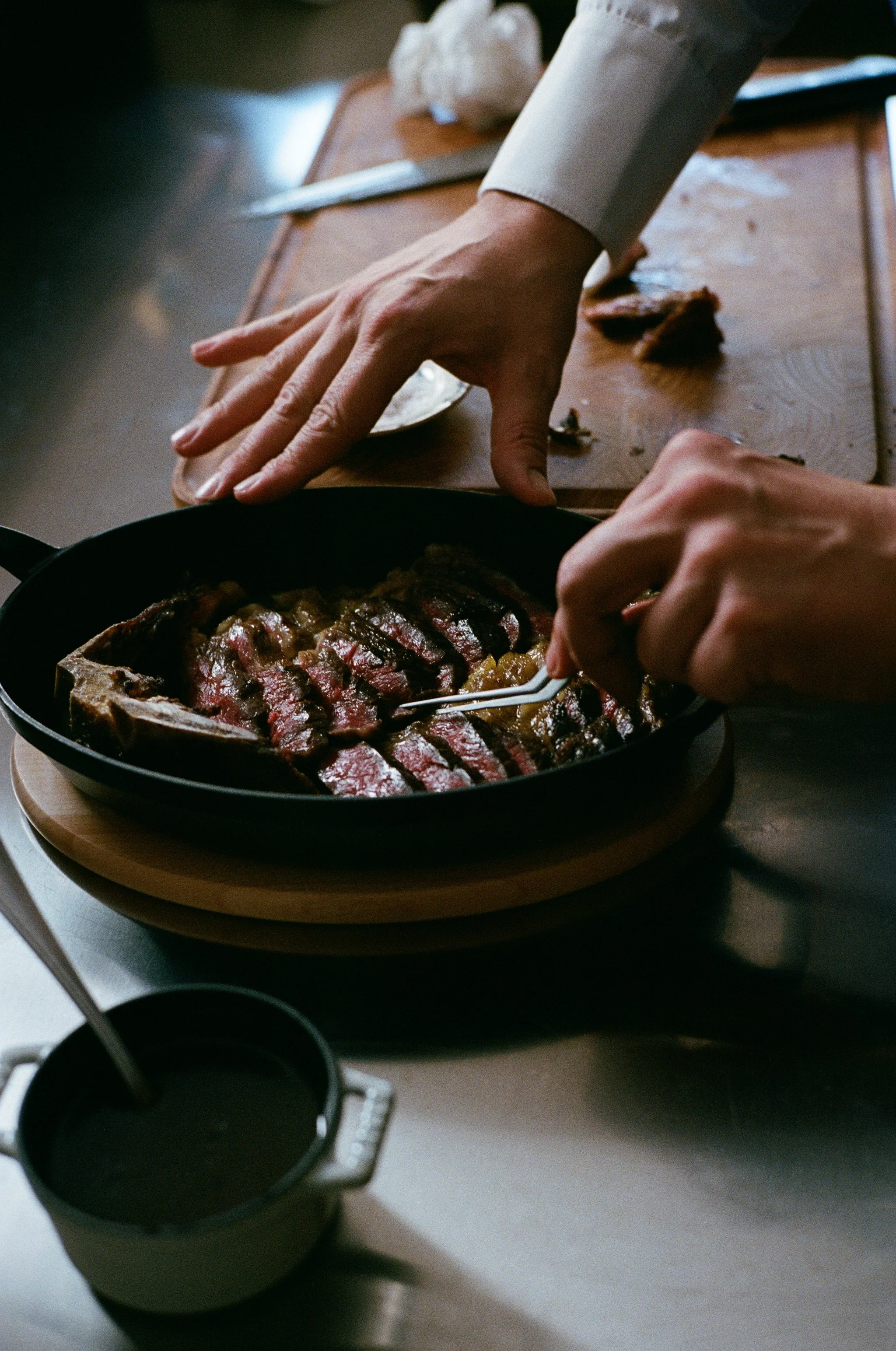 Une personne découpe un steak de bœuf cuit dans une poêle noire, avec un autre morceau de viande et des outils de cuisine sur une planche en bois.