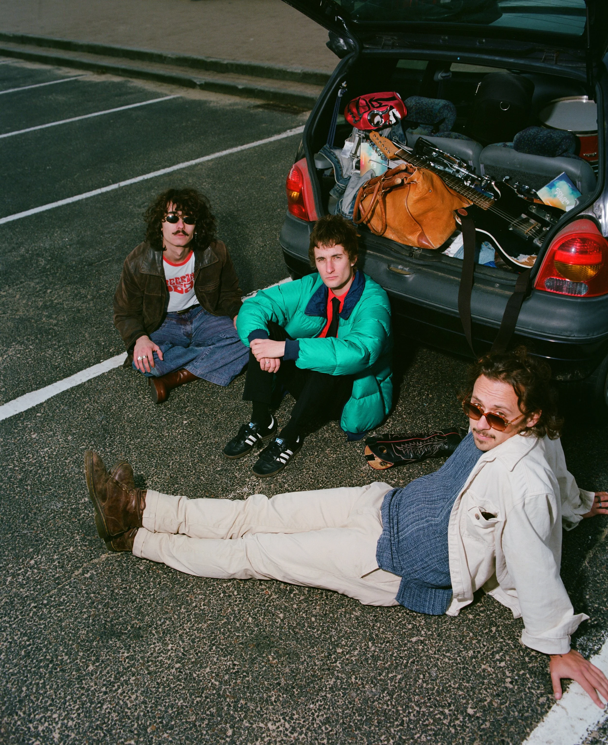 Trois jeunes hommes assis sur le sol d'un parking, devant une voiture dont le coffre est ouvert, rempli d'instruments de musique et de bagages.