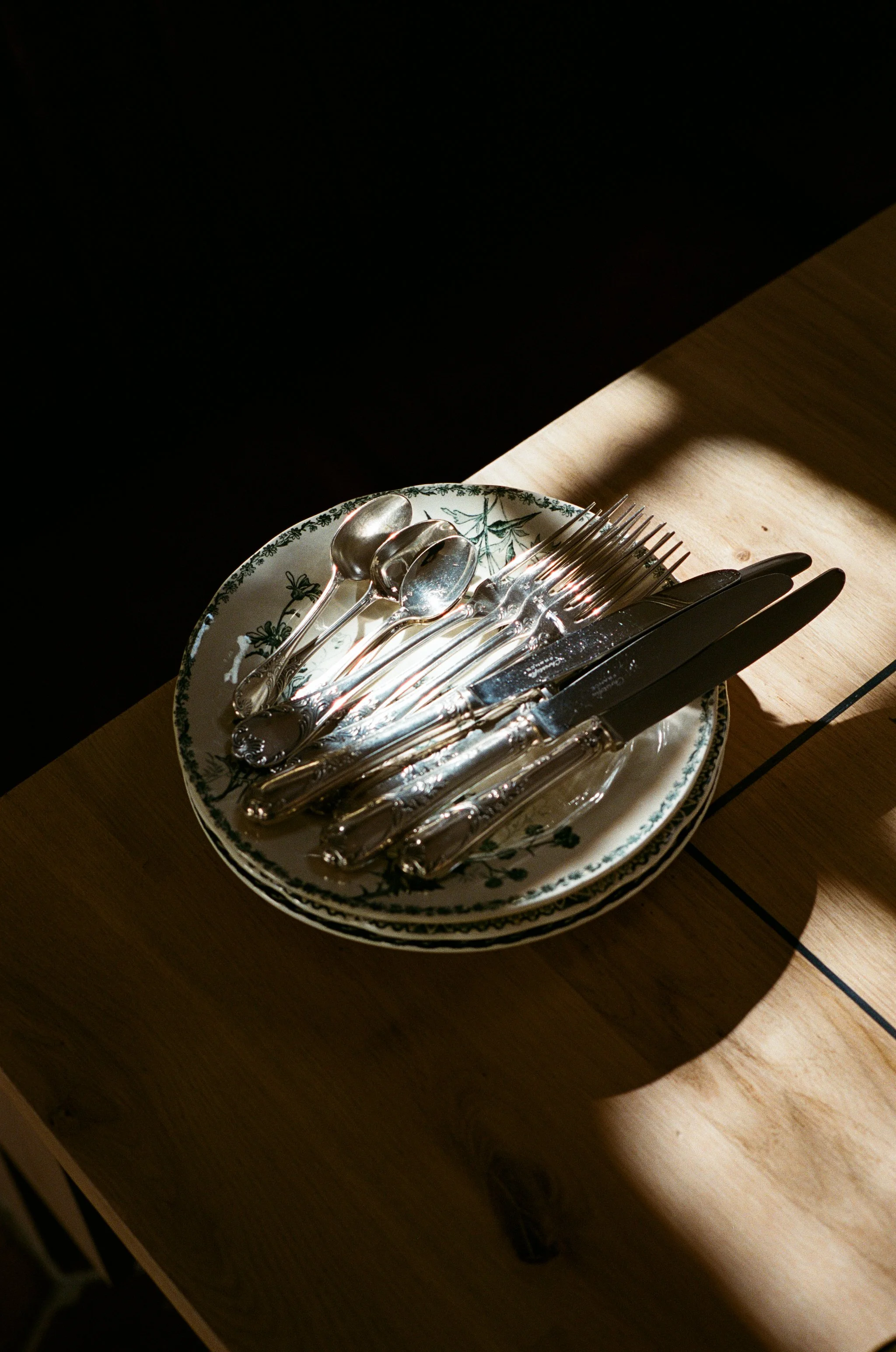 Assiette en porcelaine avec fourchettes, couteaux, cuillères en métal, positionnées sur une table en bois, avec lumière contrastée.