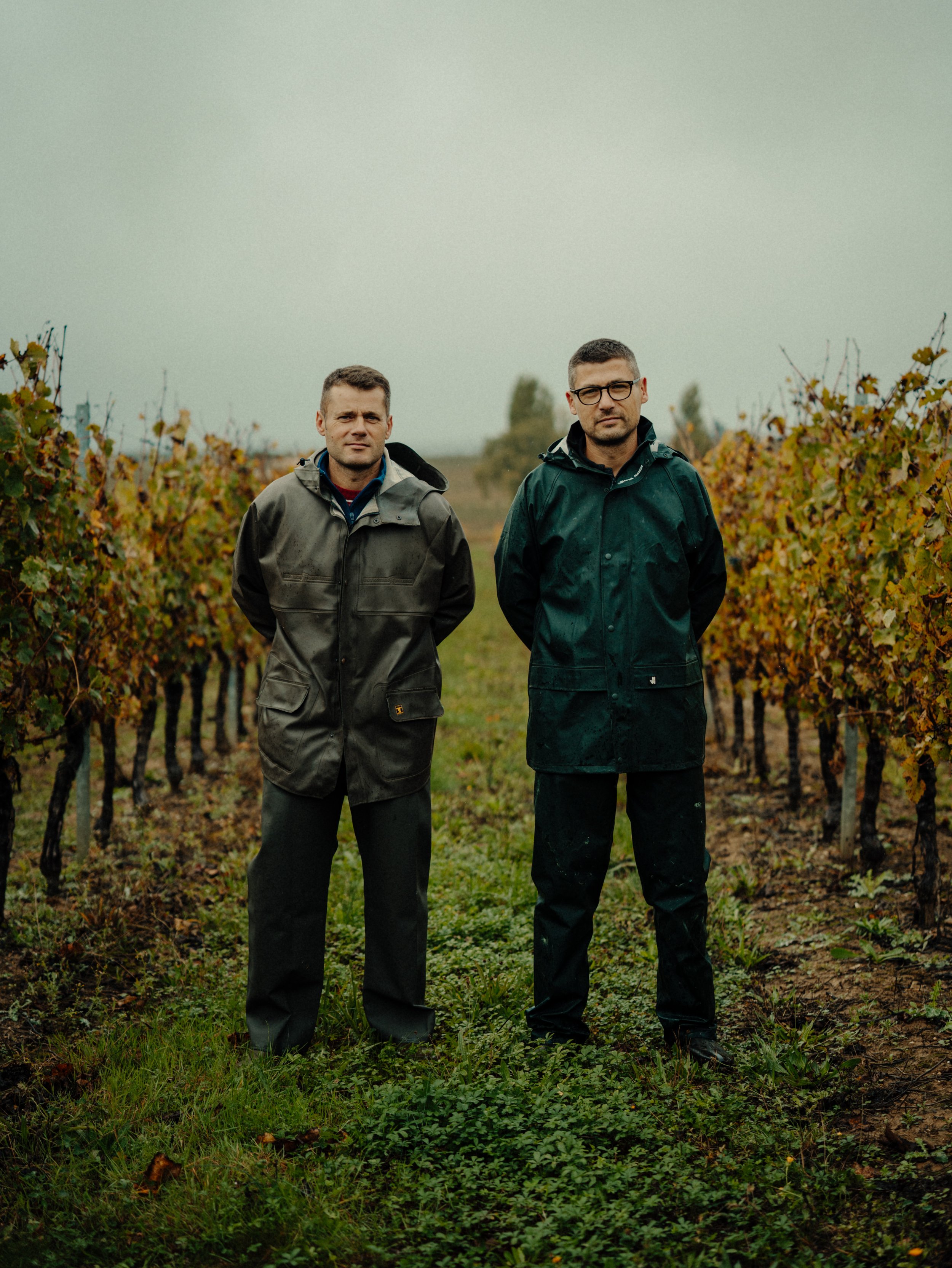 Deux hommes debout dans un vignoble sous un ciel nuageux, portant des vestes imperméables, l'un avec des lunettes