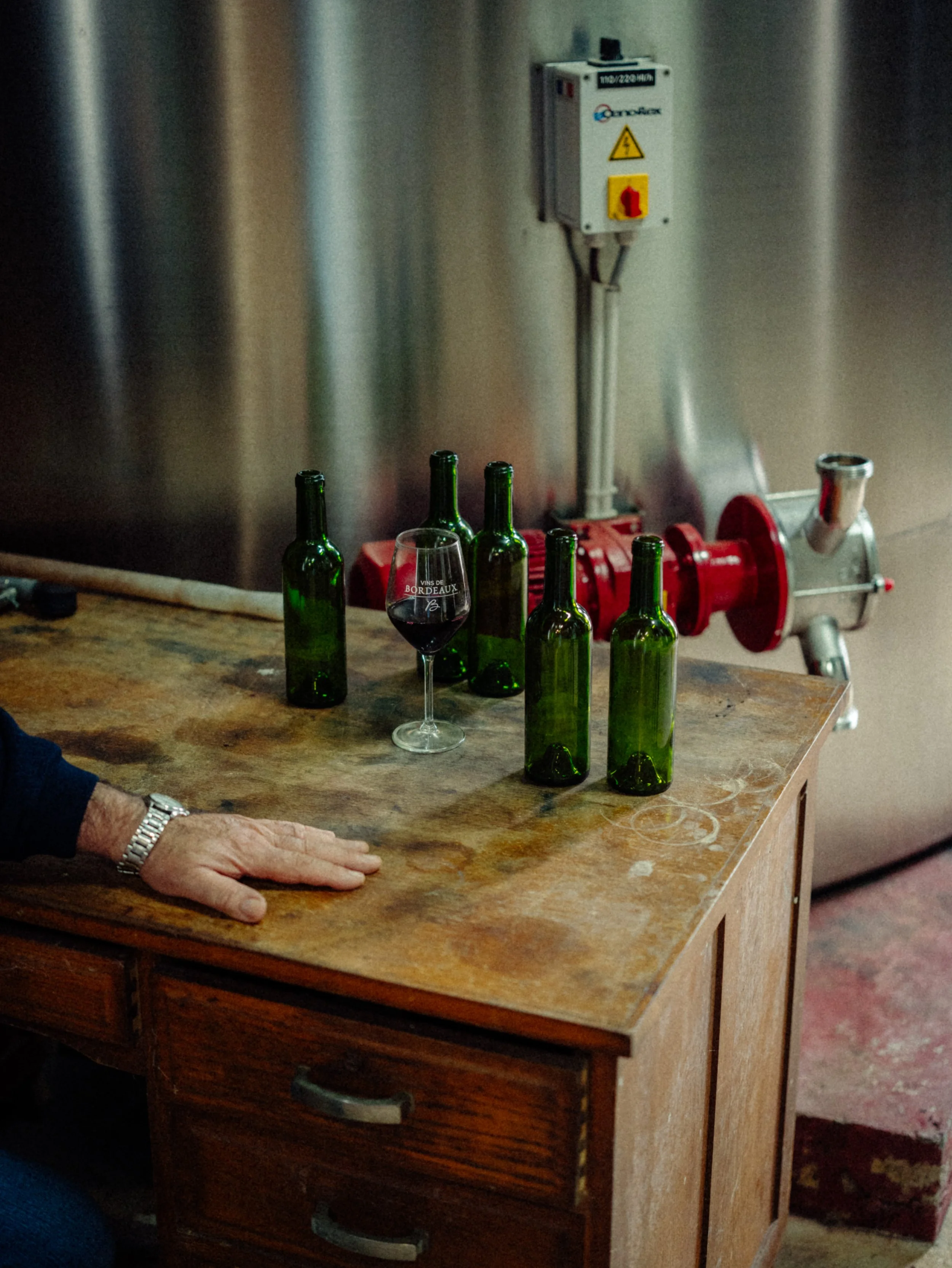 Une table en bois avec cinq bouteilles de vin vertes et un verre de vin rouge, dans un environnement industriel ou de cave à vin.