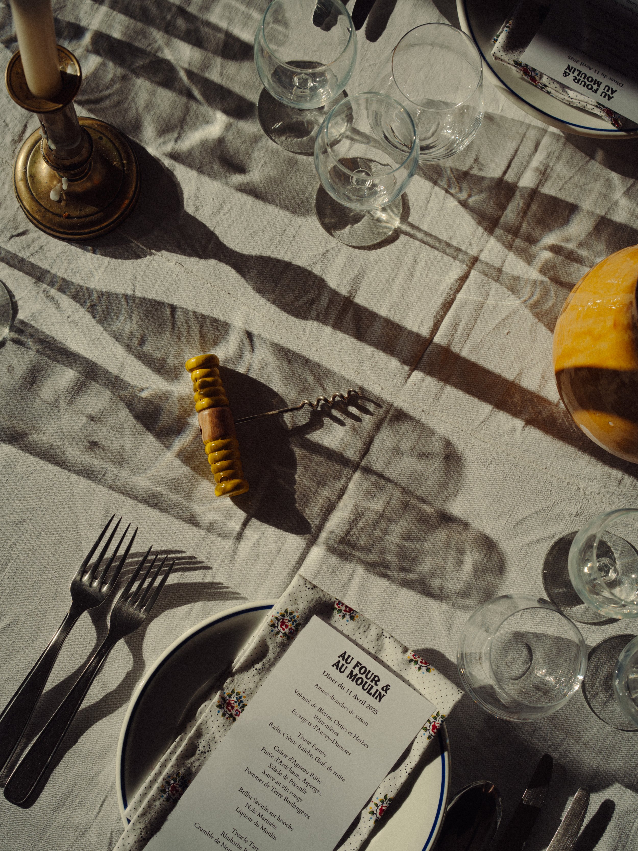 Table dressée avec des verres, une assiette, une nappe en textile, une bougie, un ouvre-bouteille, un menu, et un morceau de fromage sur la table.