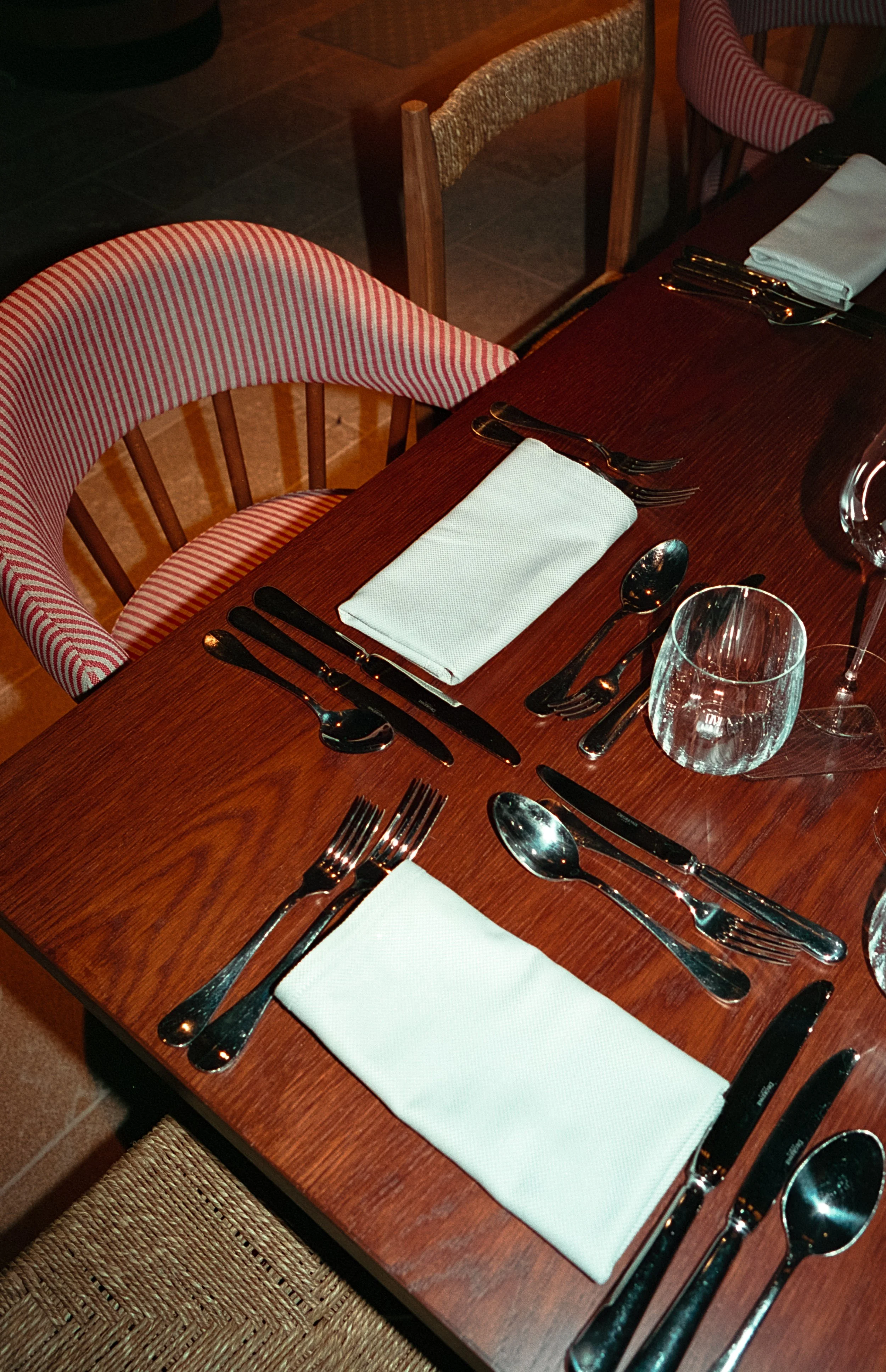 Une table de restaurant prête pour un repas, avec des couverts, des verres à vin, des serviettes blanches sur une surface en bois, et des chaises en bois avec habillage rouge rayé.