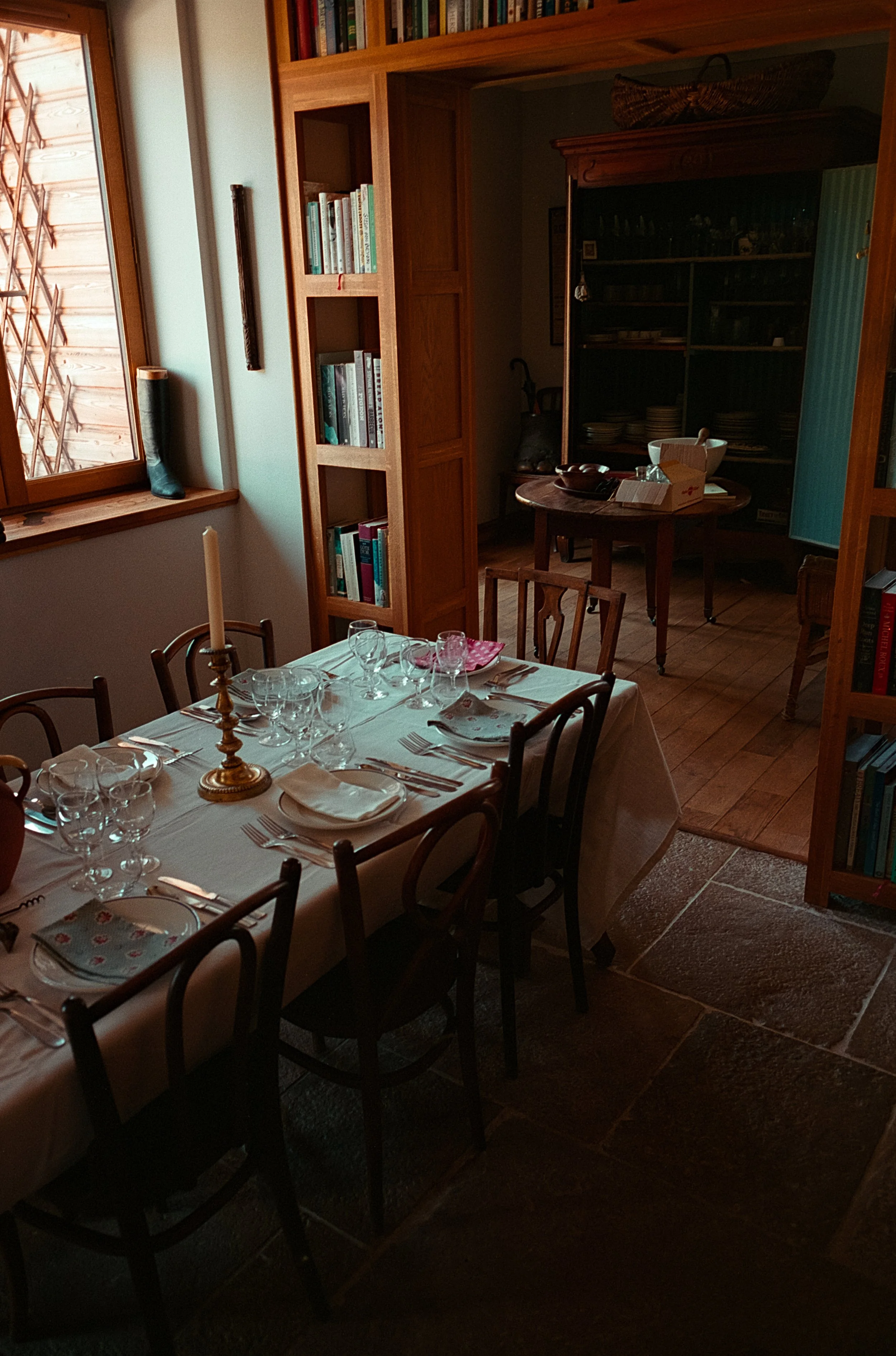 Une salle à manger chaleureuse avec une table dressée pour un repas, des verres et de la vaisselle, entourée de chaises en bois, une cheminée en arrière-plan, et une bibliothèque en bois contenant des livres.