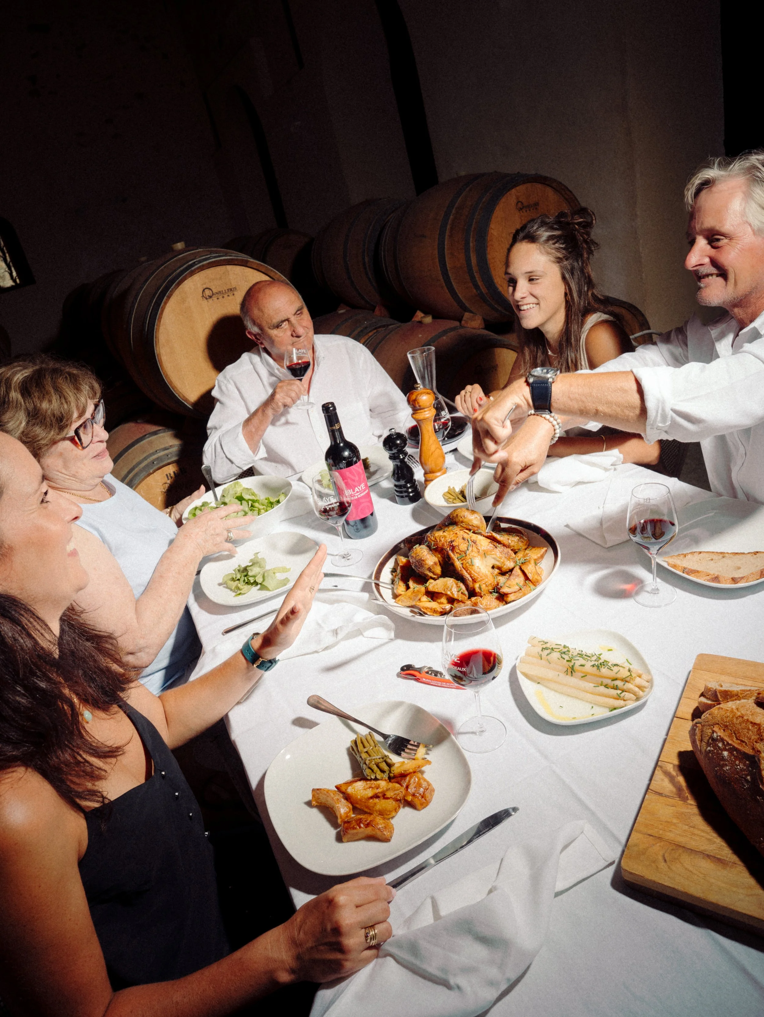 Groupe de personnes autour d'une table à manger un dîner avec une cuisine raffinée, à l'intérieur d'une cave à vin, avec de grands tonneaux en bois en arrière-plan.
