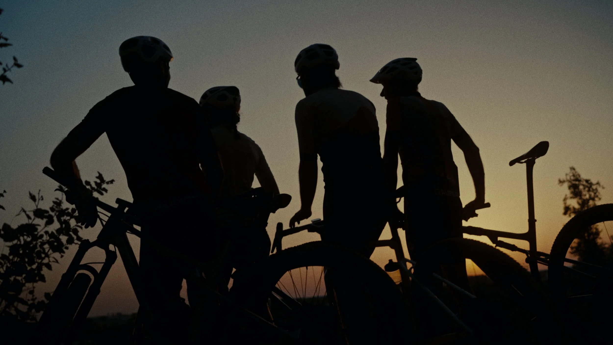 Silhouettes de cinq cyclistes portant des casques, debout avec leurs vélos au coucher de soleil.