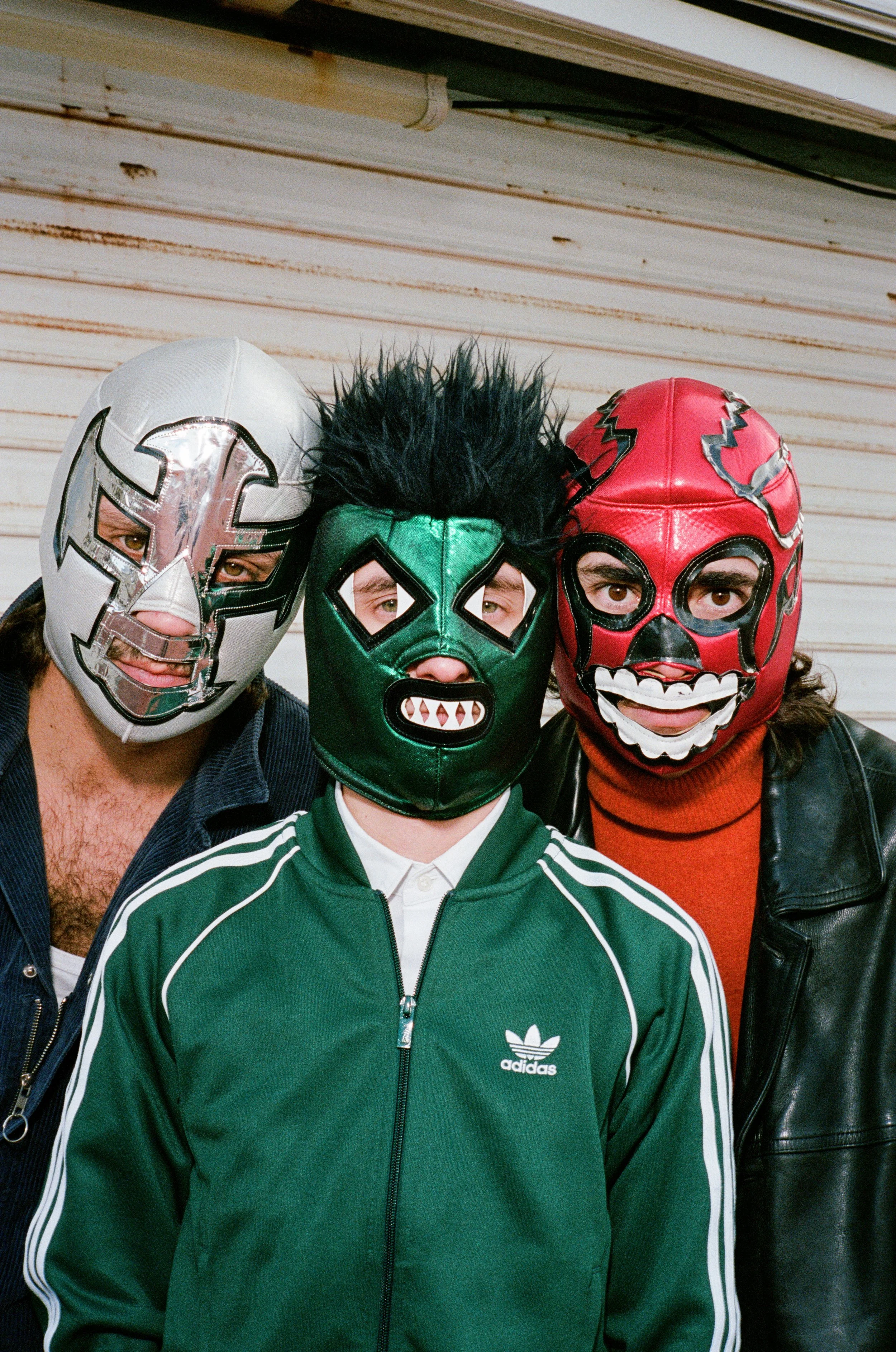 Trois personnes portant des masques de luchador colorés et un t-shirt Adidas vert, posant devant une porte en métal rouillé.