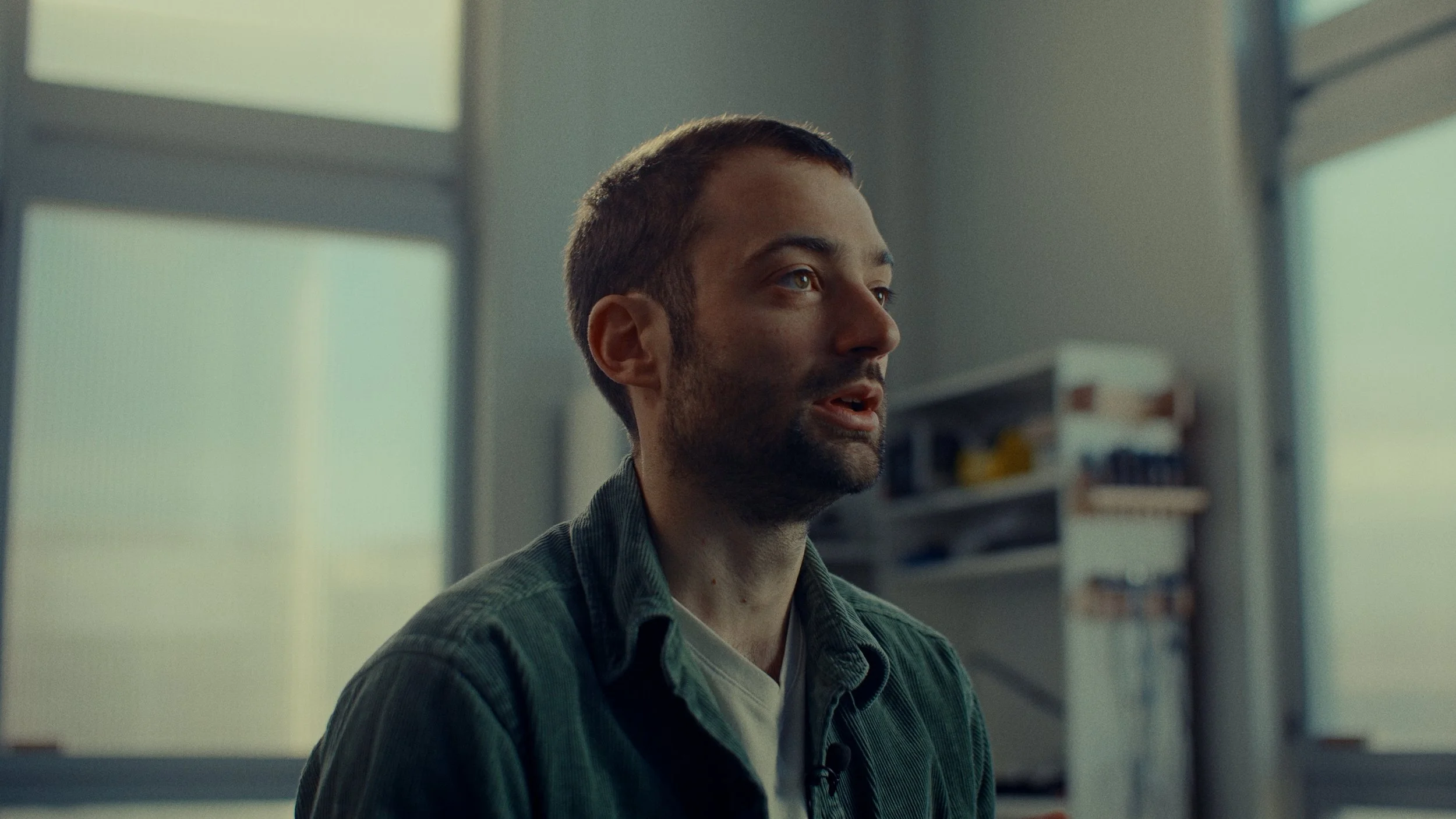 Homme avec barbe, cheveux courts, portant une chemise en jeans, assis dans une pièce avec grandes fenêtres et étagère en arrière-plan.