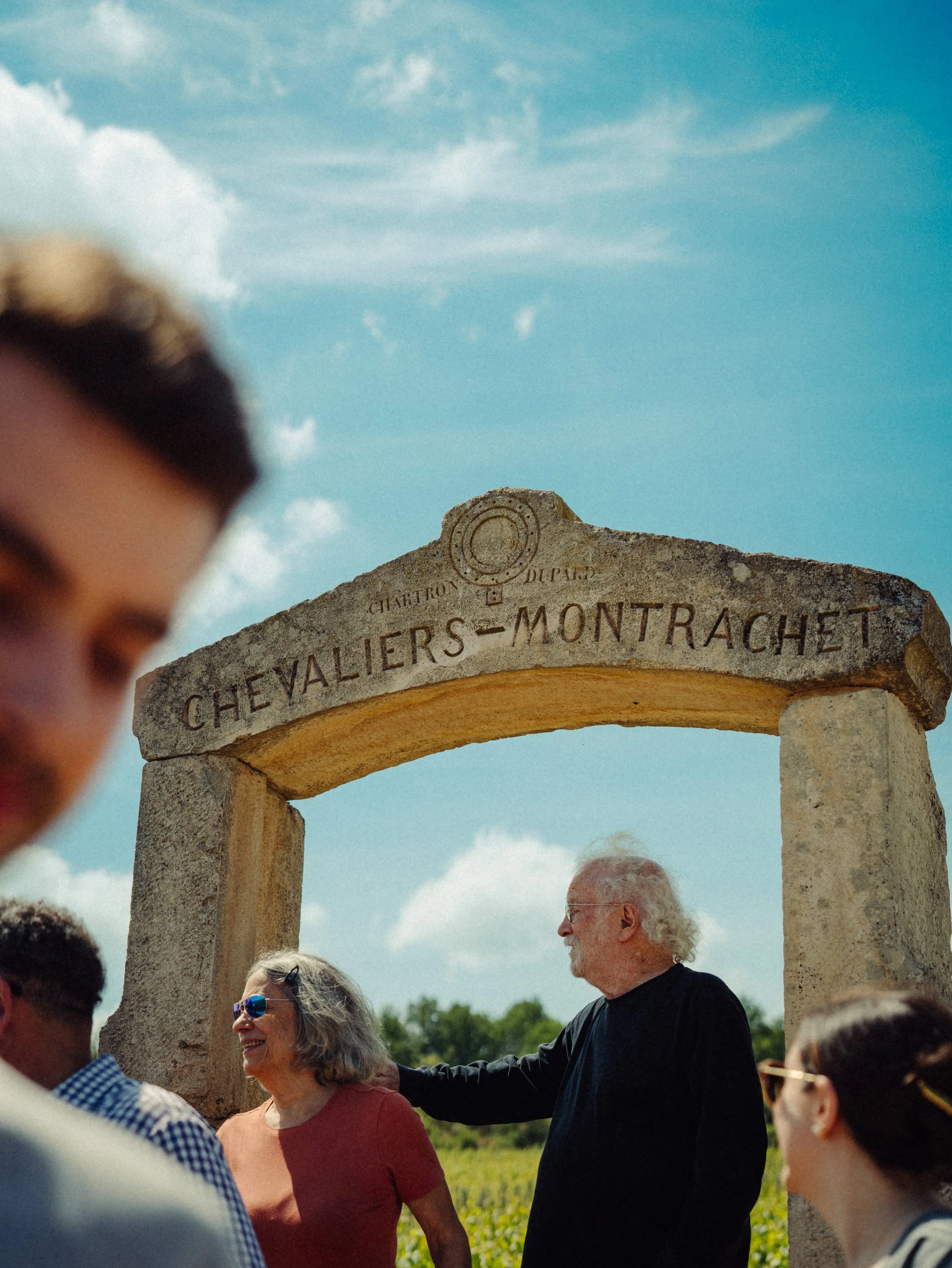 Groupe de personnes devant une arche en pierre avec l'inscription "Chevaliers-Montrachet", sous un ciel bleu avec quelques nuages.