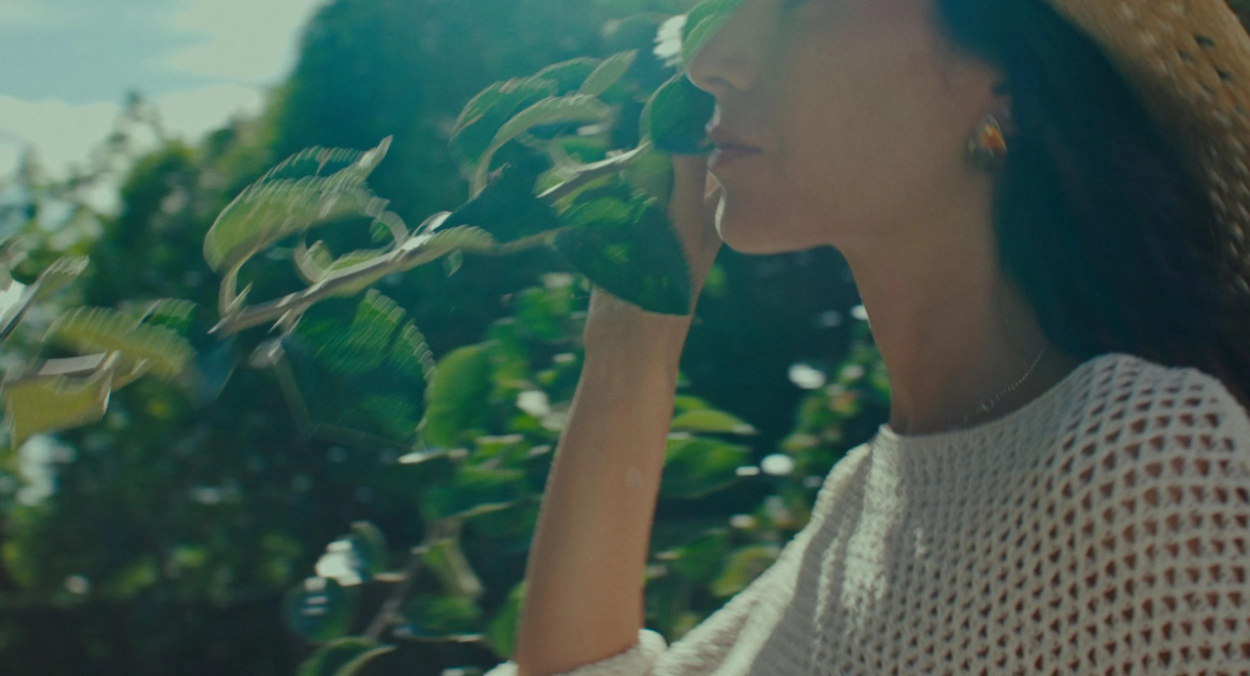 Femme avec boucle d'oreilles, portant un pull en crochet blanc, touchant une branche d'arbre en plein air lors d'une journée ensoleillée.