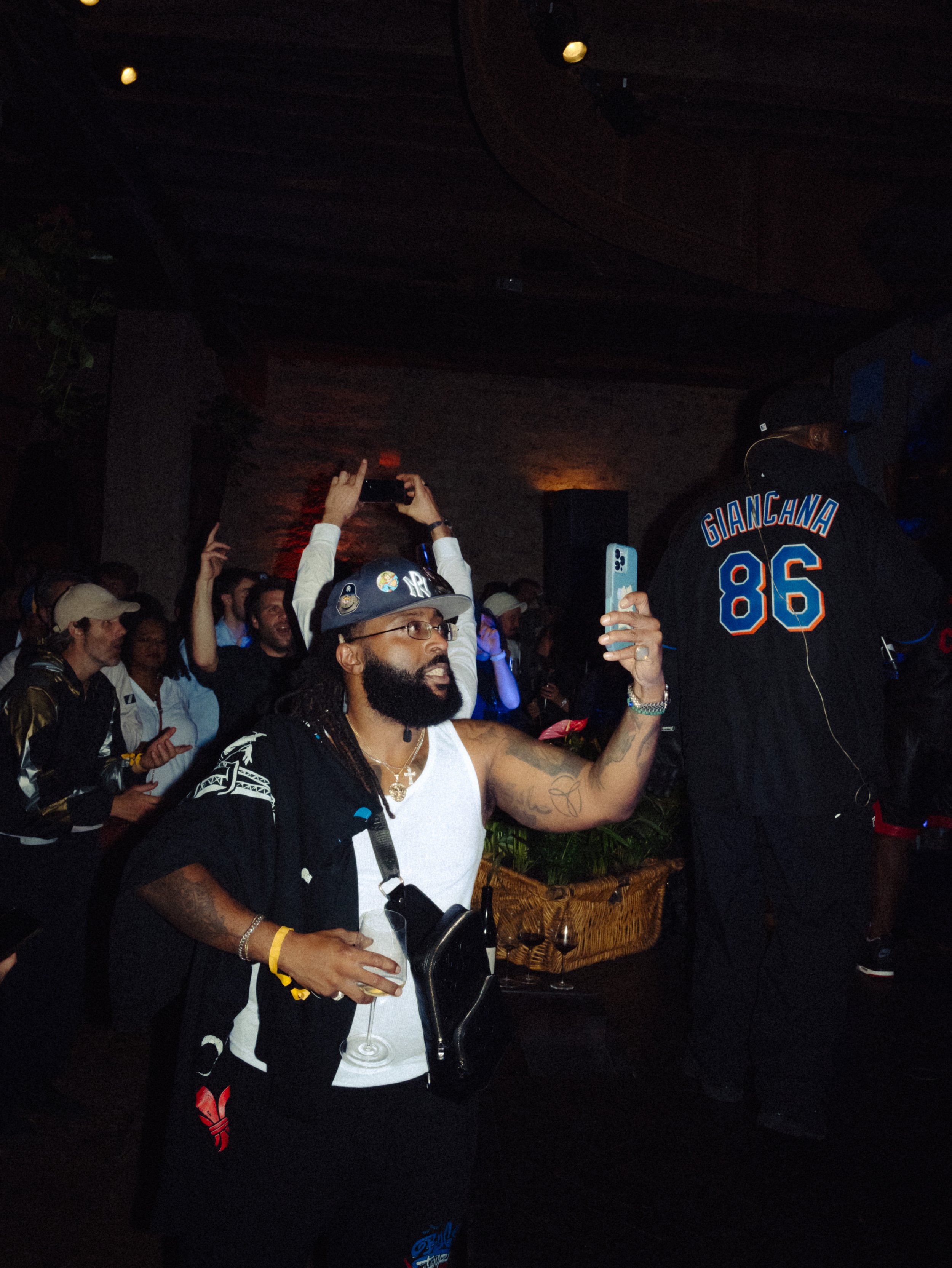 Homme avec dreadlocks, lunettes et veste noire prenant une selfie dans une fête ou un événement, entouré d'autres personnes qui s'amusent.