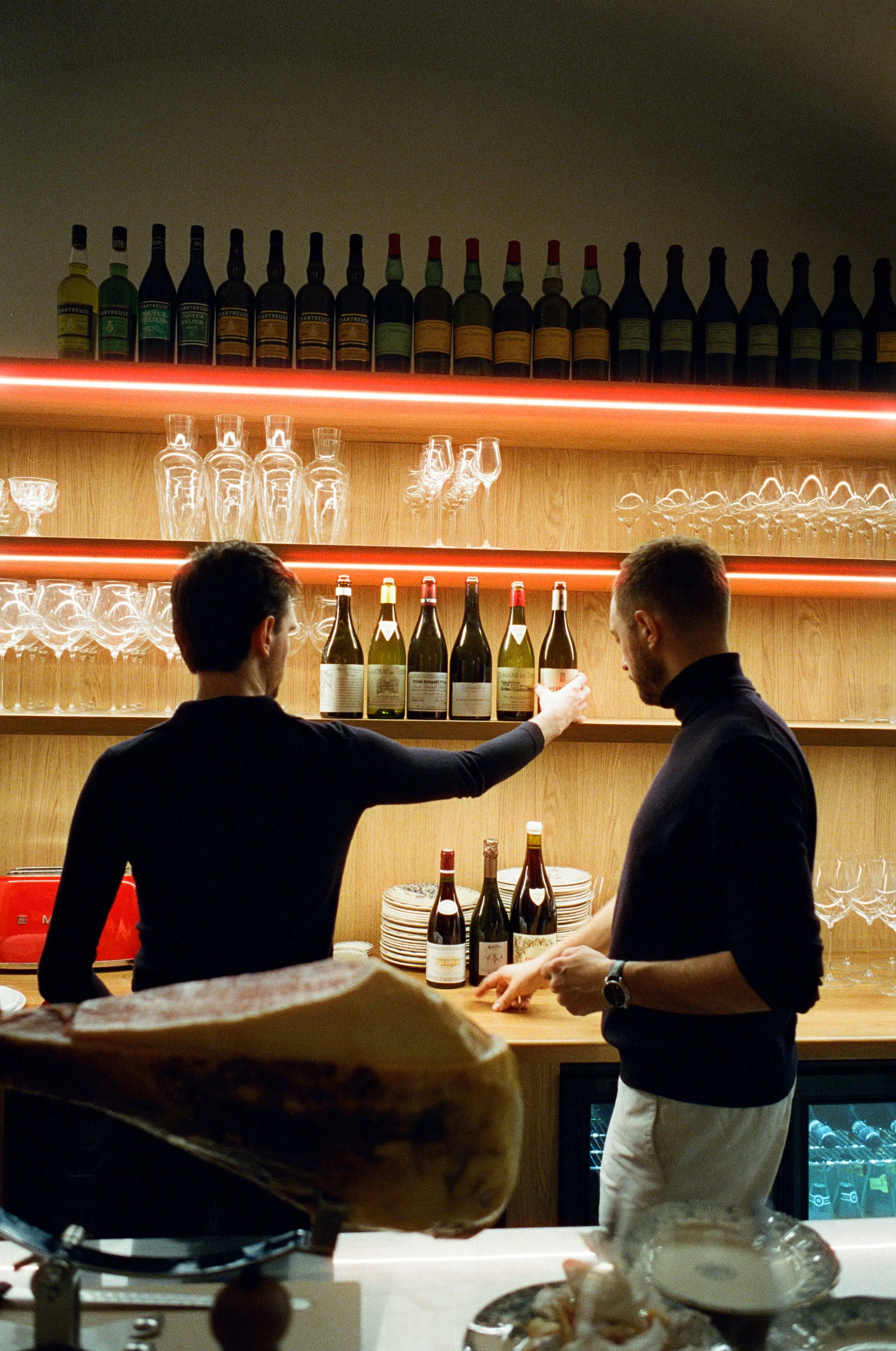 Deux personnes discutent au bar, avec des bouteilles de vin et des verres derrière eux, dans un décor chaleureux en bois.