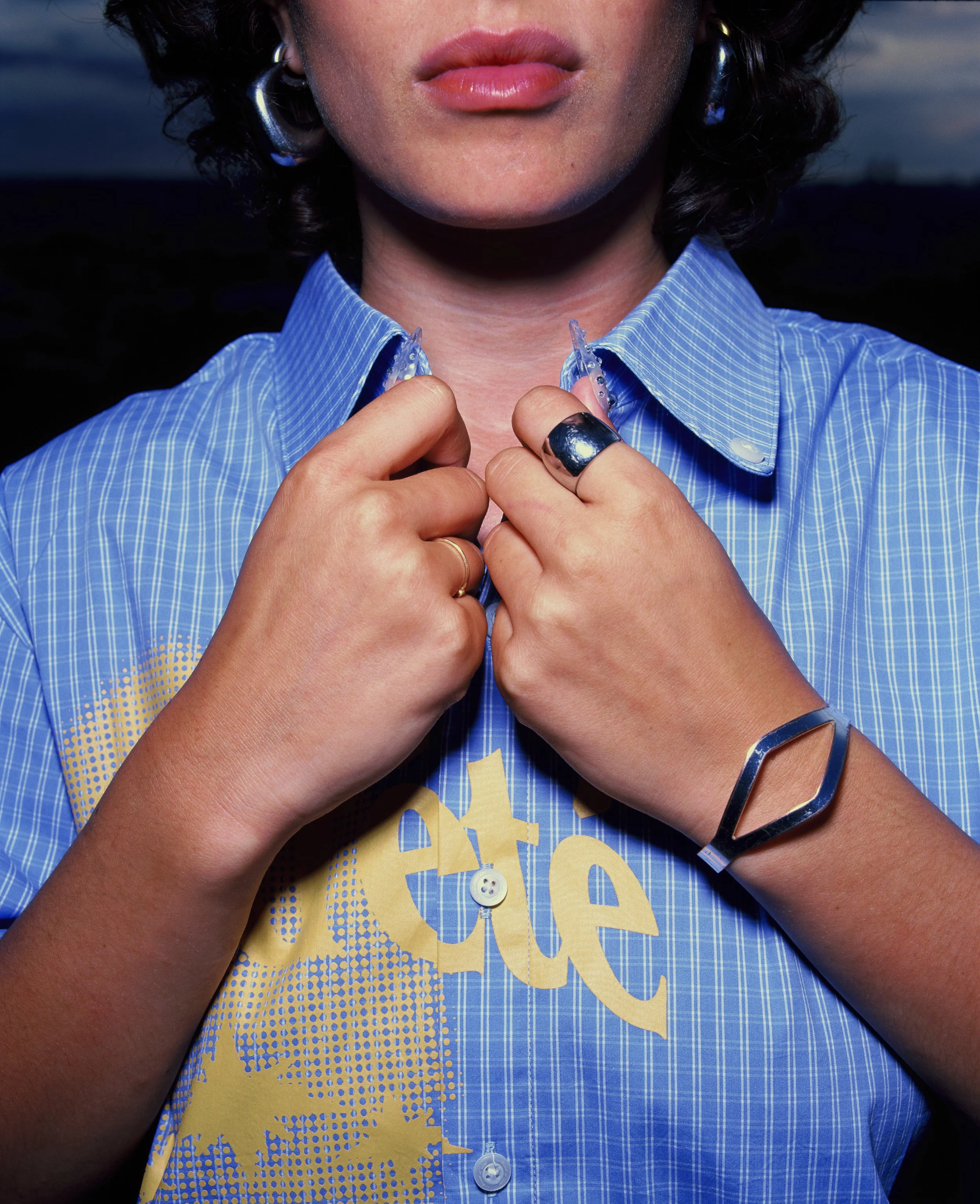 Une femme portant une chemise bleue avec un motif jaune, ajustant le col. Elle porte des accessoires en métal, notamment une bague, un bracelet en forme de losange et une boucle d'oreille.