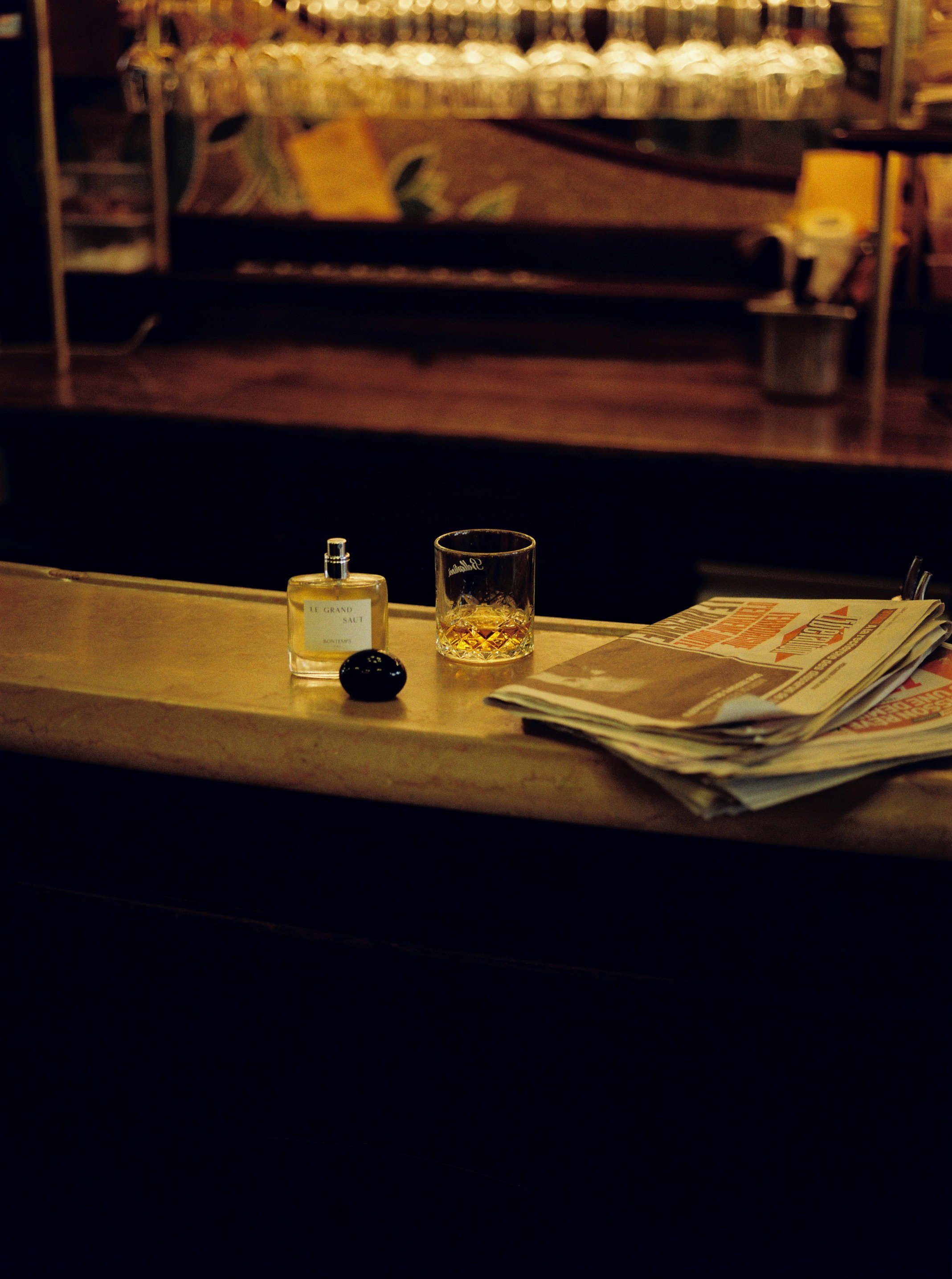 Une table de bar avec un flacon de parfum, un verre à whisky avec une boisson, et un journal.